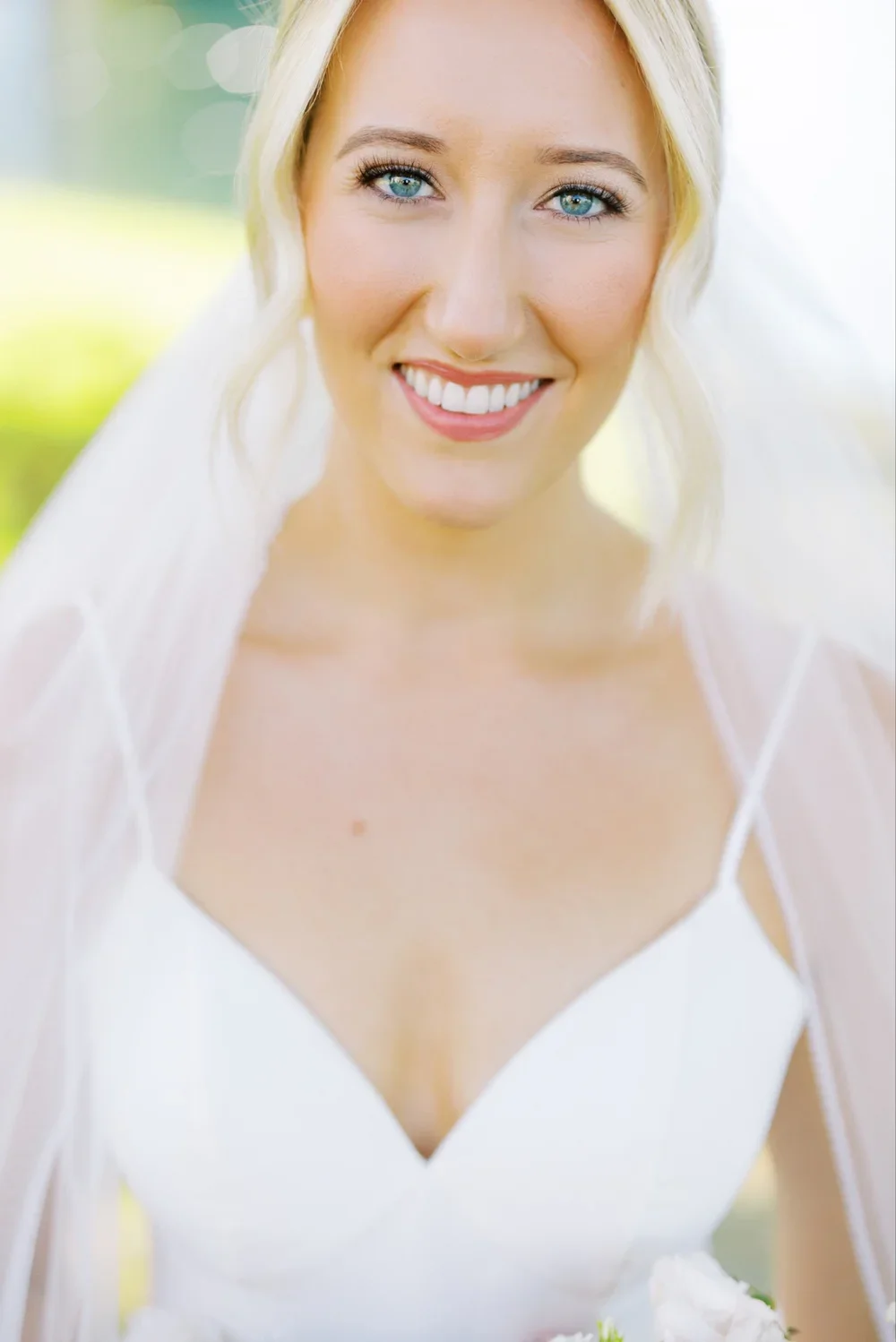A smiling bride with blonde hair, blue eyes, wearing a white wedding dress with spaghetti straps, outdoors with a blurred green background.