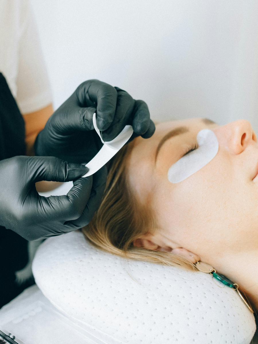 A woman receiving a facial treatment, with a skincare professional applying a product to her face while her eyes are closed, wearing a white eye patch.