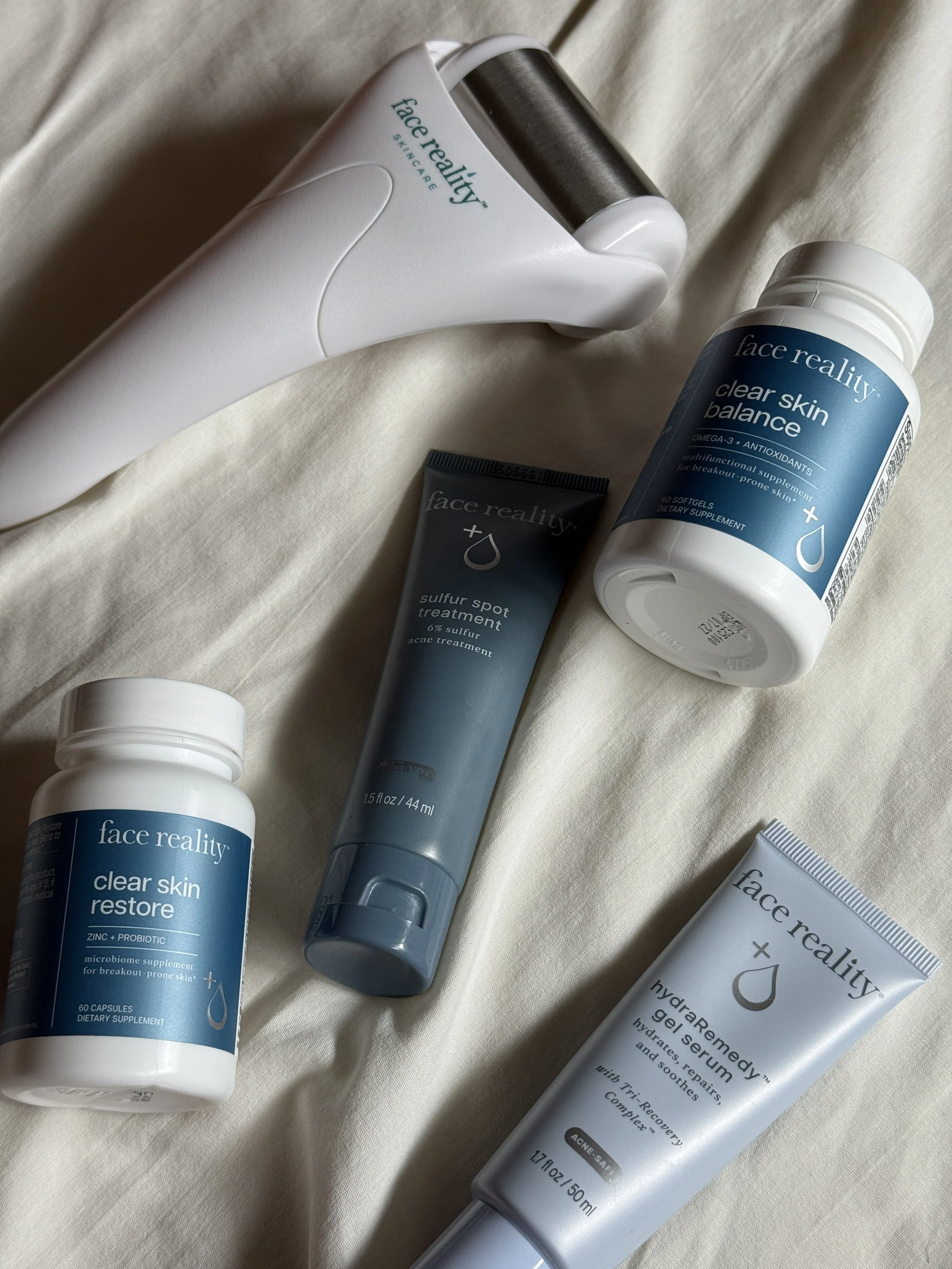 Various skincare and supplement products from face reality, including a facial roller, a capsule bottle, a softur spot treatment tube, and a gel serum tube, arranged on a light-colored fabric surface.