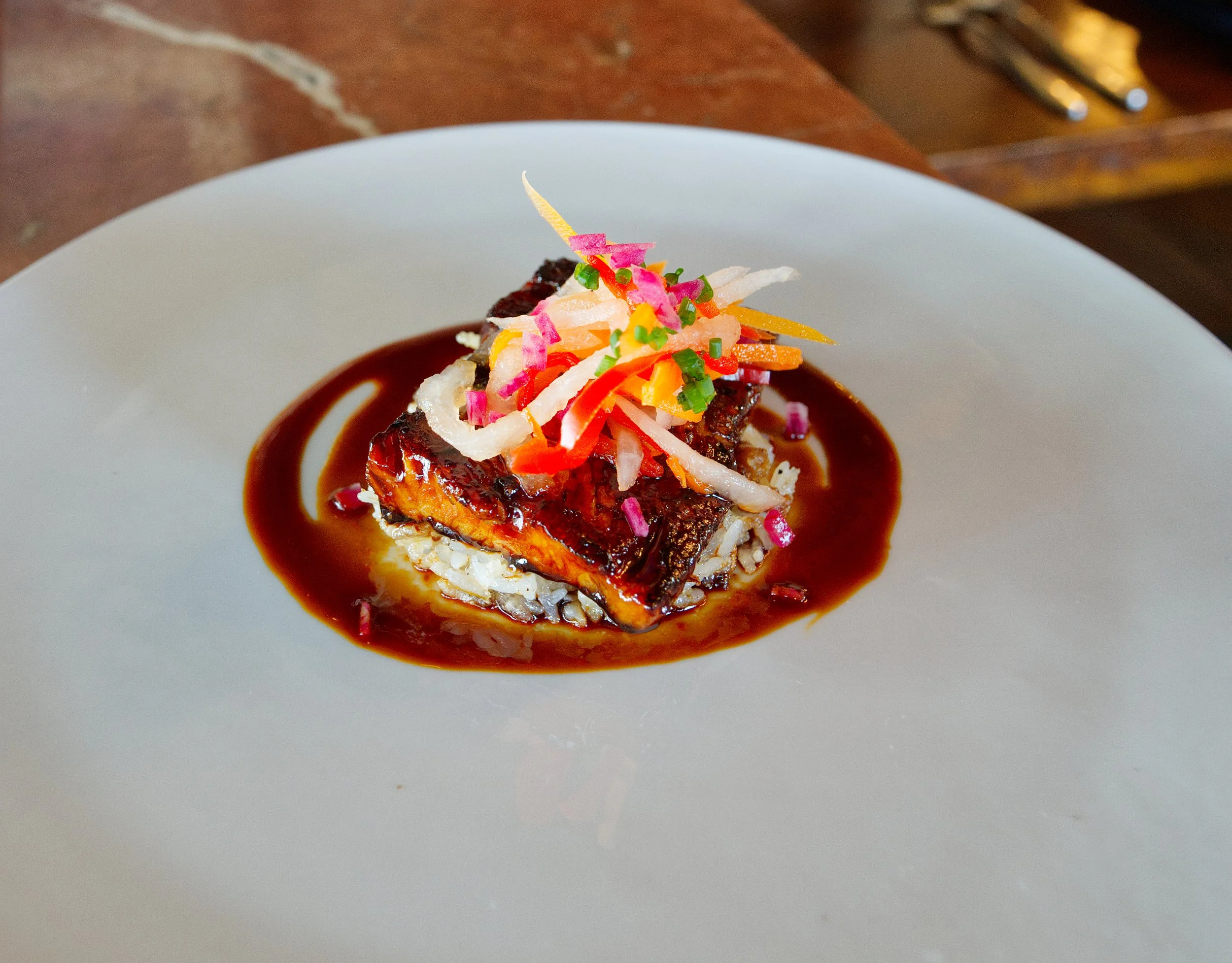 A plated dish featuring a piece of meat topped with shredded vegetables and colorful garnishes, surrounded by a dark sauce on a white plate.