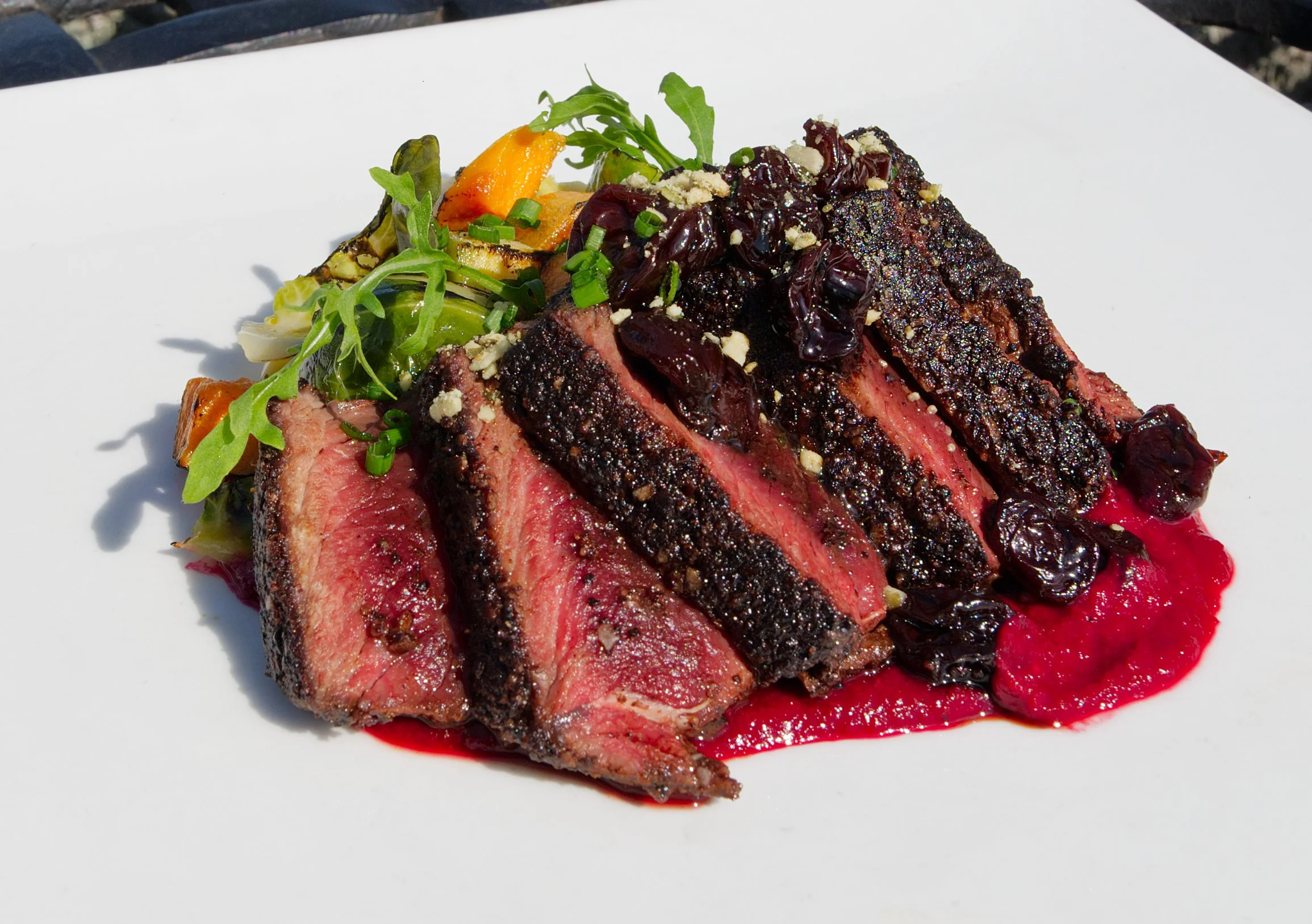 Sliced medium-rare steak topped with a dark berry sauce, garnished with chopped nuts, green herbs, and accompanied by grilled vegetables on a white plate.