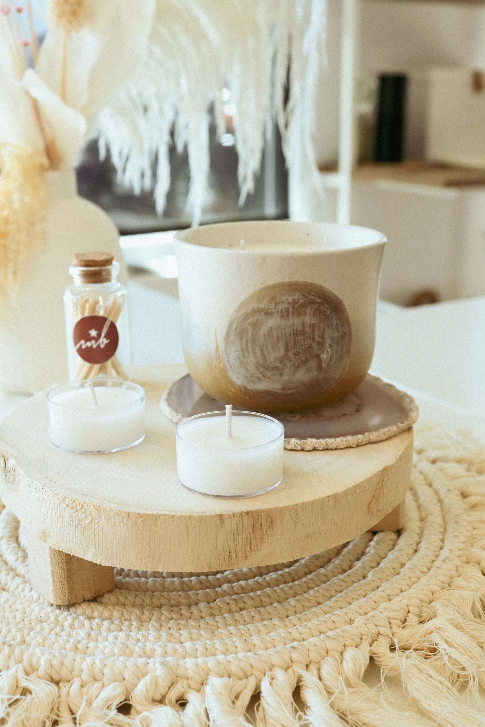 Decorative centerpiece with candles, small jar, and a large ceramic bowl on a wooden slab, placed on a woven mat in a cozy room.