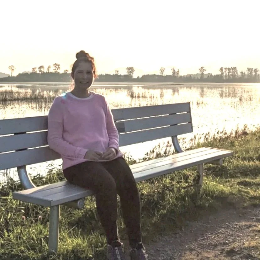 A woman sitting on a white park bench near a body of water during sunset, with trees in the background.