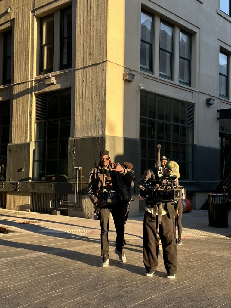Two crew members filming with cameras on a city street in front of a building, with the sun casting long shadows