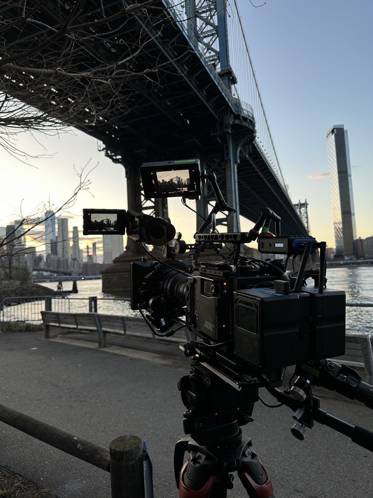 Professional film camera set up outdoors near the water, with the Manhattan Bridge overhead and city skyline in the background at sunset.