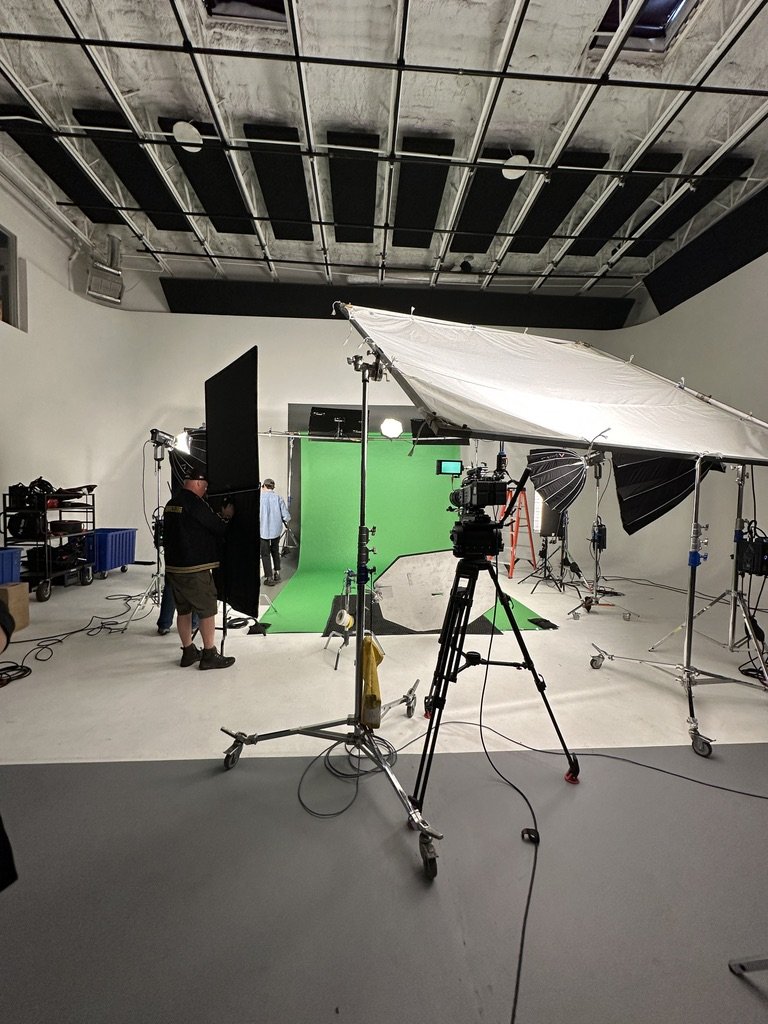 A professional photography or video production studio with a green screen backdrop, lighting equipment, cameras, and crew setting up for filming.