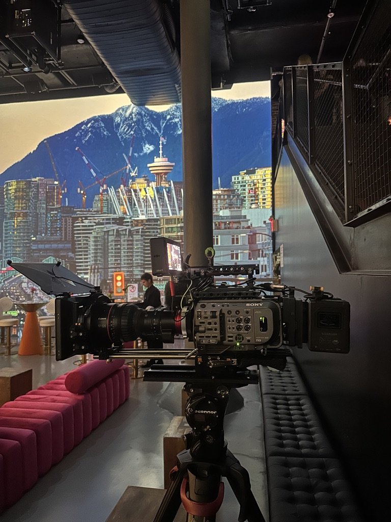 View of a professional video camera on a tripod inside a modern building, filming a cityscape backdrop with tall buildings, some under construction, and mountainous terrain in the background.