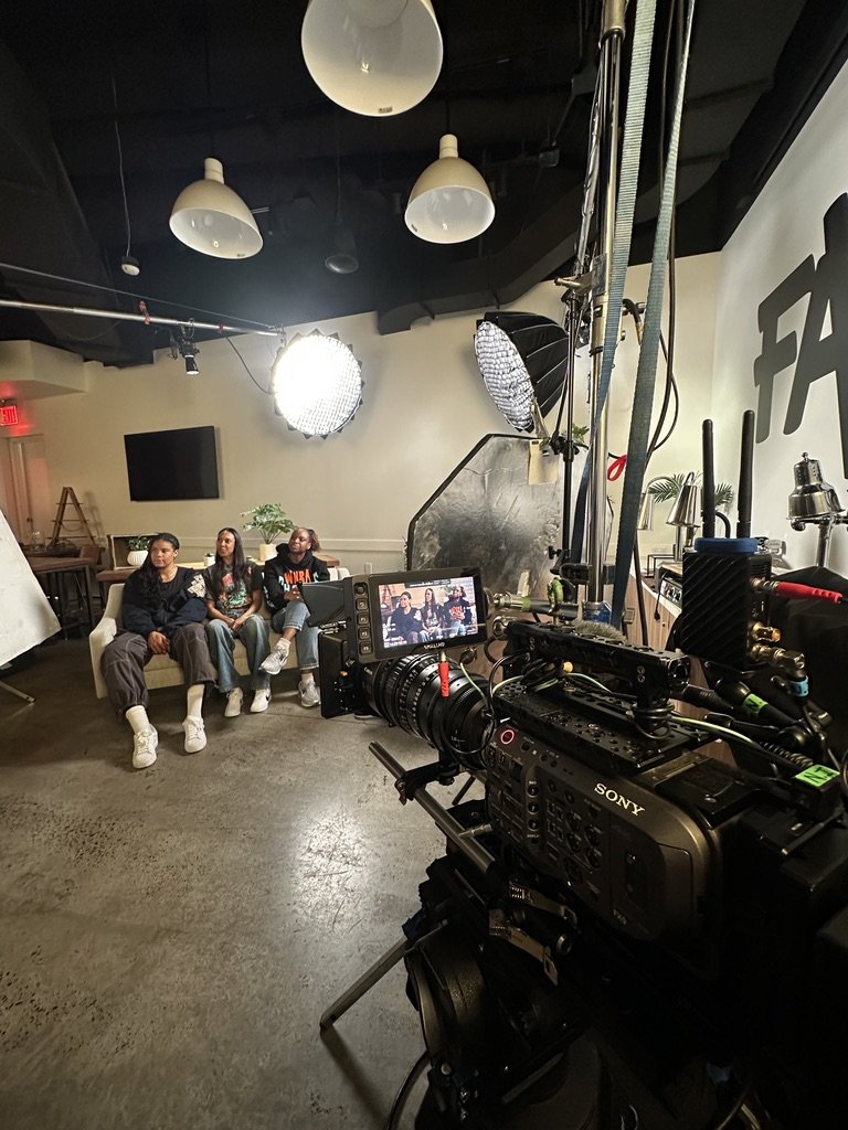 Behind the scenes of a video shoot with three young women sitting on a couch, studio lighting equipment, a Sony camera on a tripod, and a white wall with partial text visible.