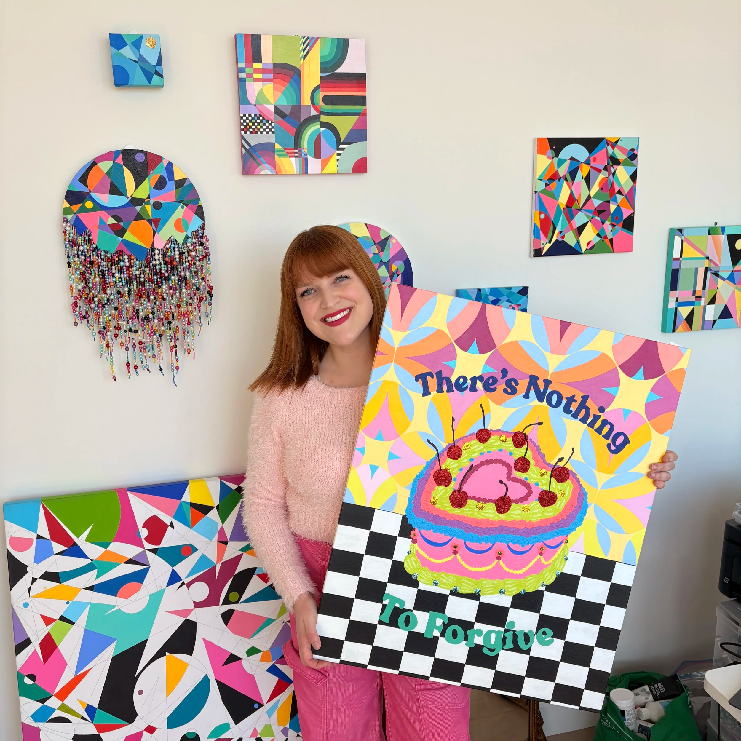 A woman with red hair and a pink sweater holding a colorful panel with a birthday cake illustration, the words "There's Nothing To Forgive" on it, in an art studio with colorful abstract paintings on the wall.