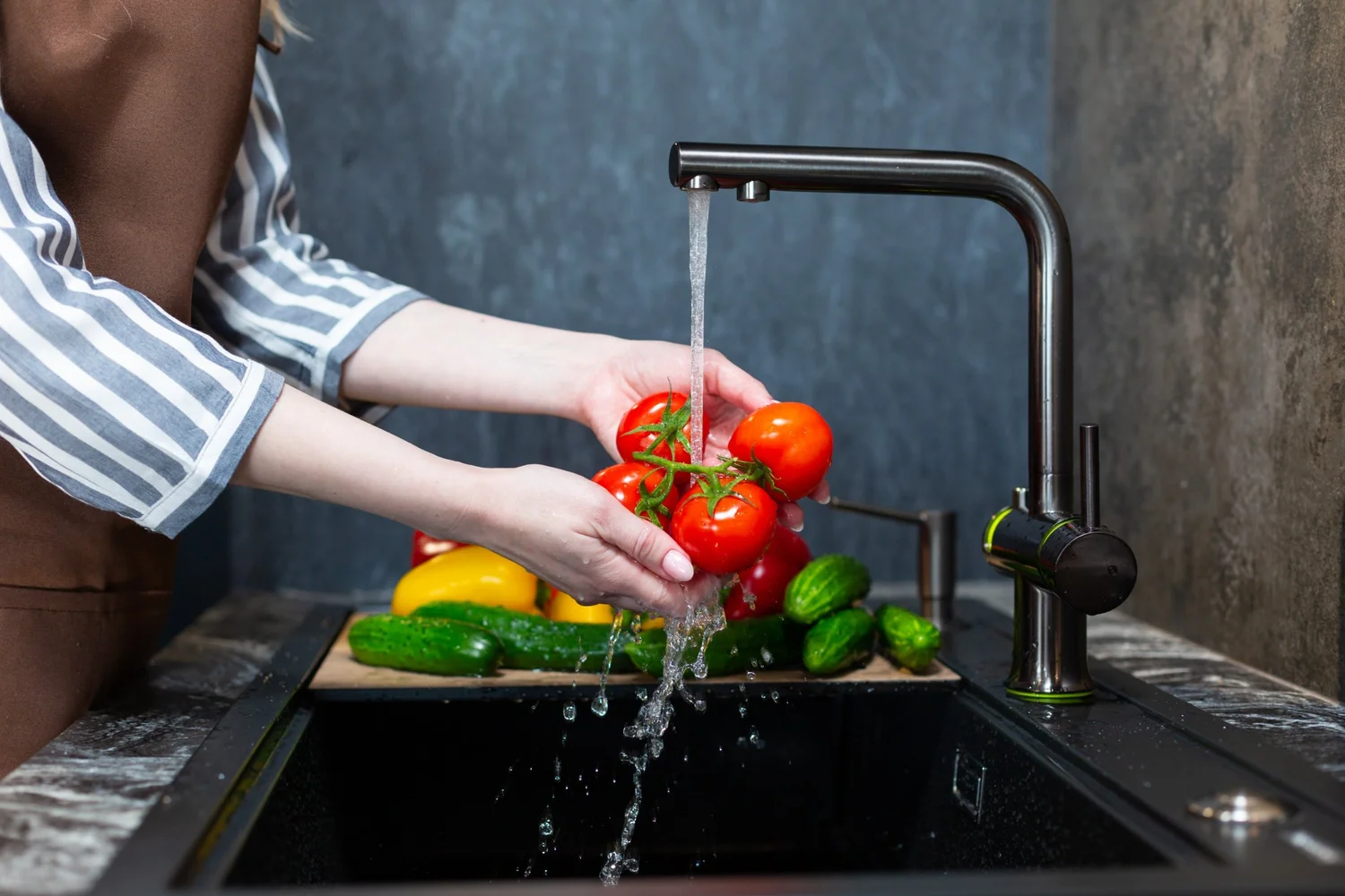 Person washing fresh tomatoes and cucumbers in a kitchen sink with running water.
