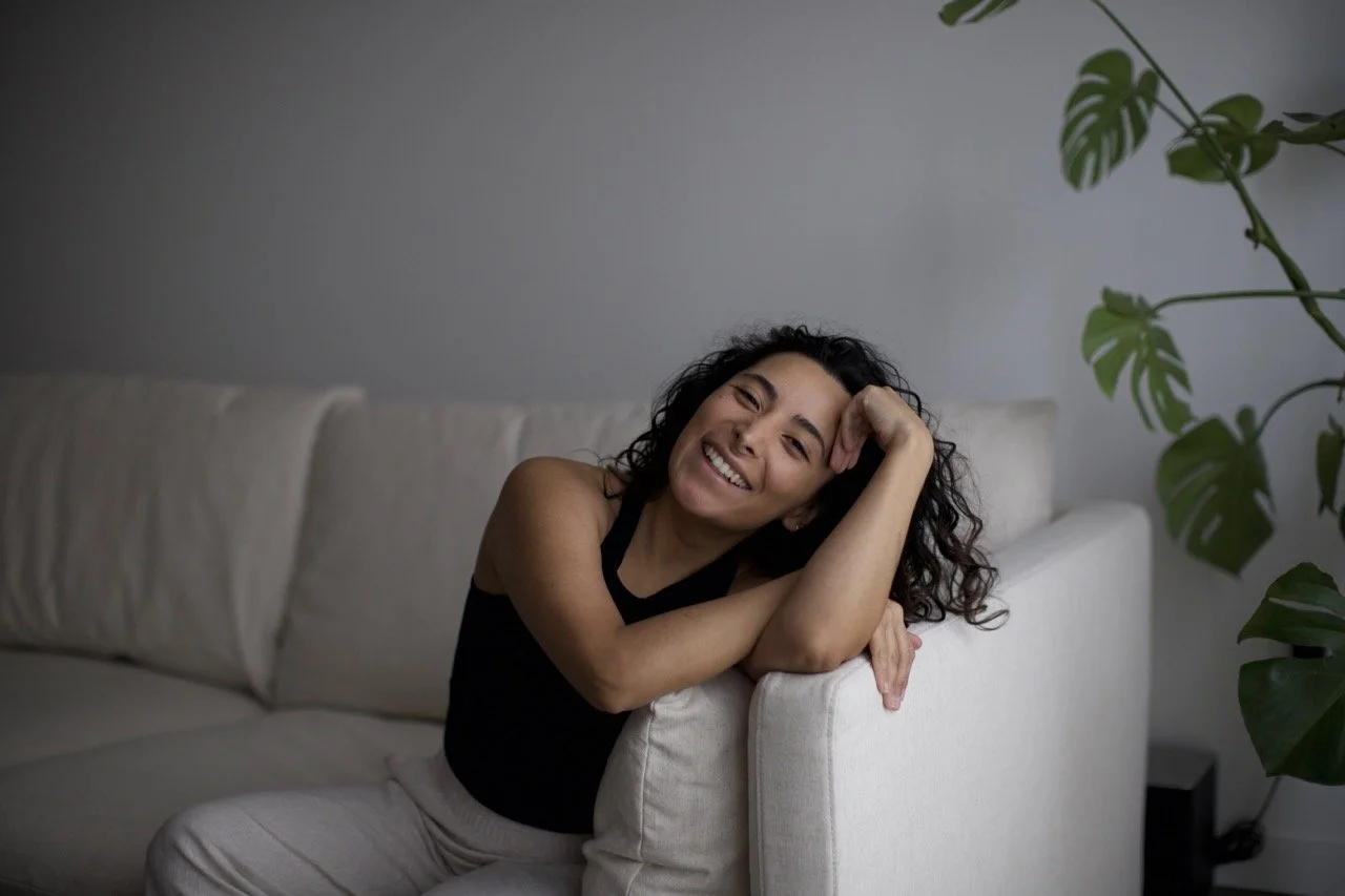 A woman with curly dark hair smiling while leaning on the arm of a light-colored couch in a room with a large green plant nearby.