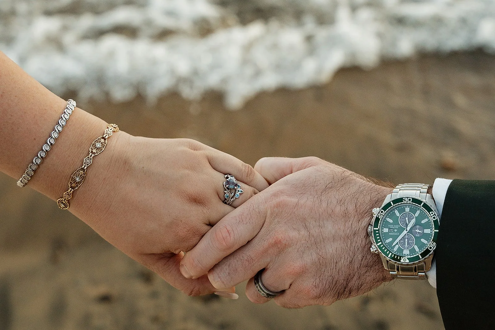 Close-up of a couple holding hands on King's Beach in Tahoe, wearing wedding rings and jewelry, with waves in the background.