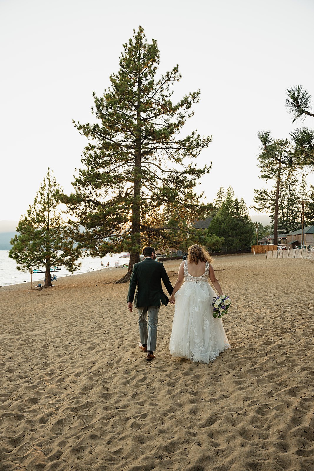A bride and groom walking on a sandy beach in Tahoe at sunset, holding hands, with trees and houses in the background.