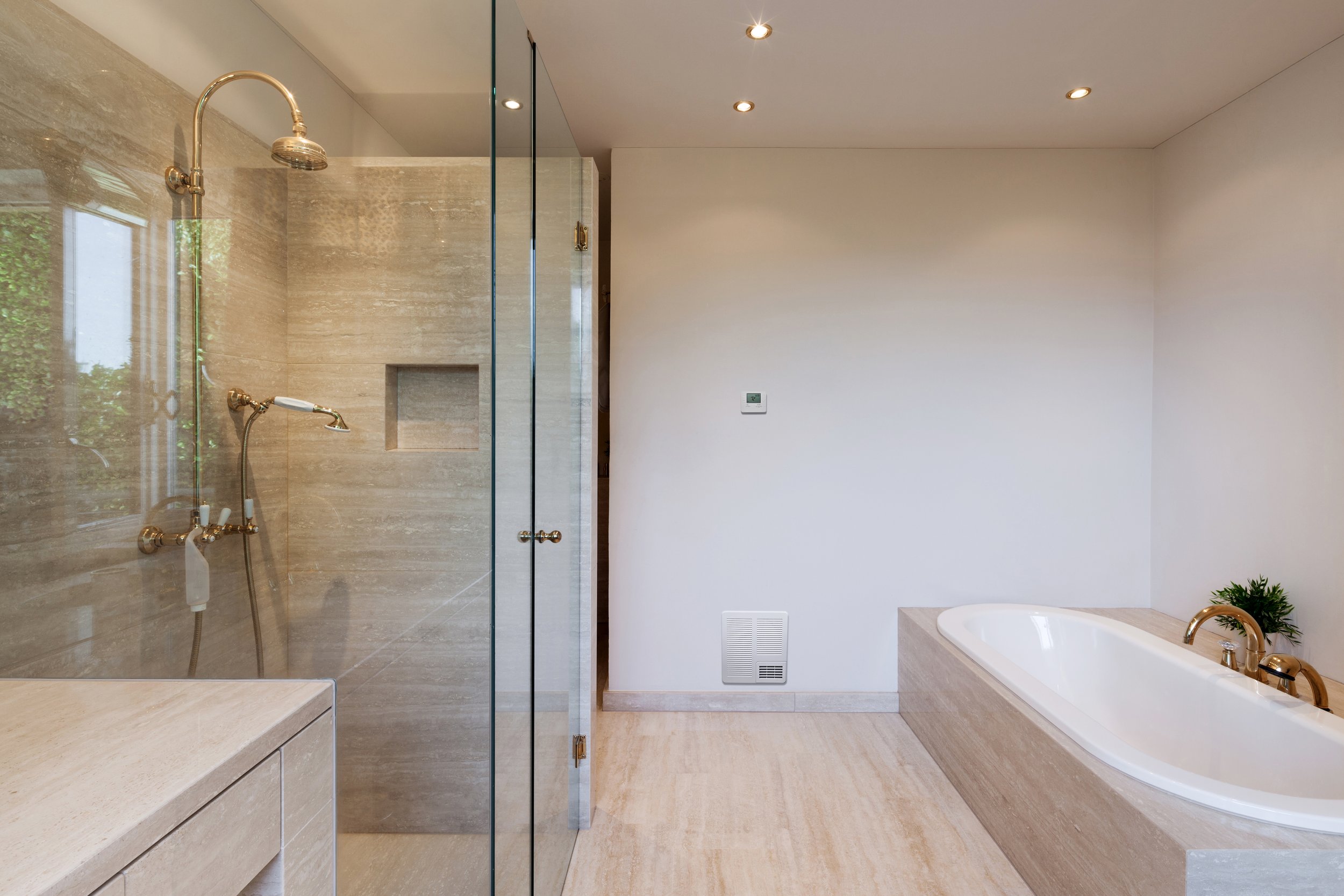 Modern bathroom with an ASH Innovations built-in wall heater. It has a glass-enclosed shower area, beige marble tiles, a white bathtub, and a small potted plant near the bathtub. Recessed ceiling lights provide illumination.