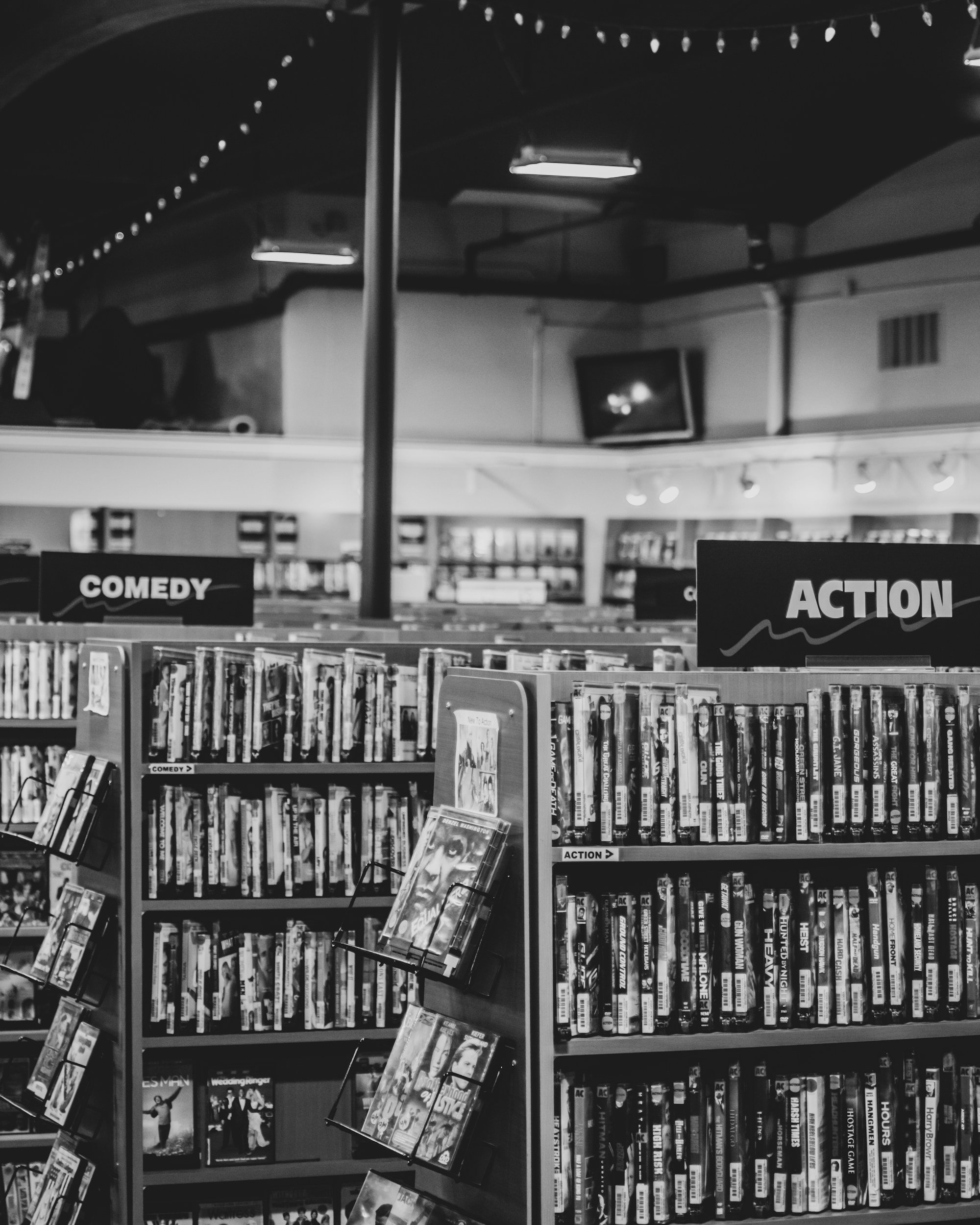 A section of a bookstore with shelves filled with books. Two signs labeled 'COMEDY' and 'ACTION' are visible above the shelves. There are DVD or book displays with titles, some partially visible, and a TV or monitor mounted on the wall in the background. The ceiling has string lights and fluorescent lighting.