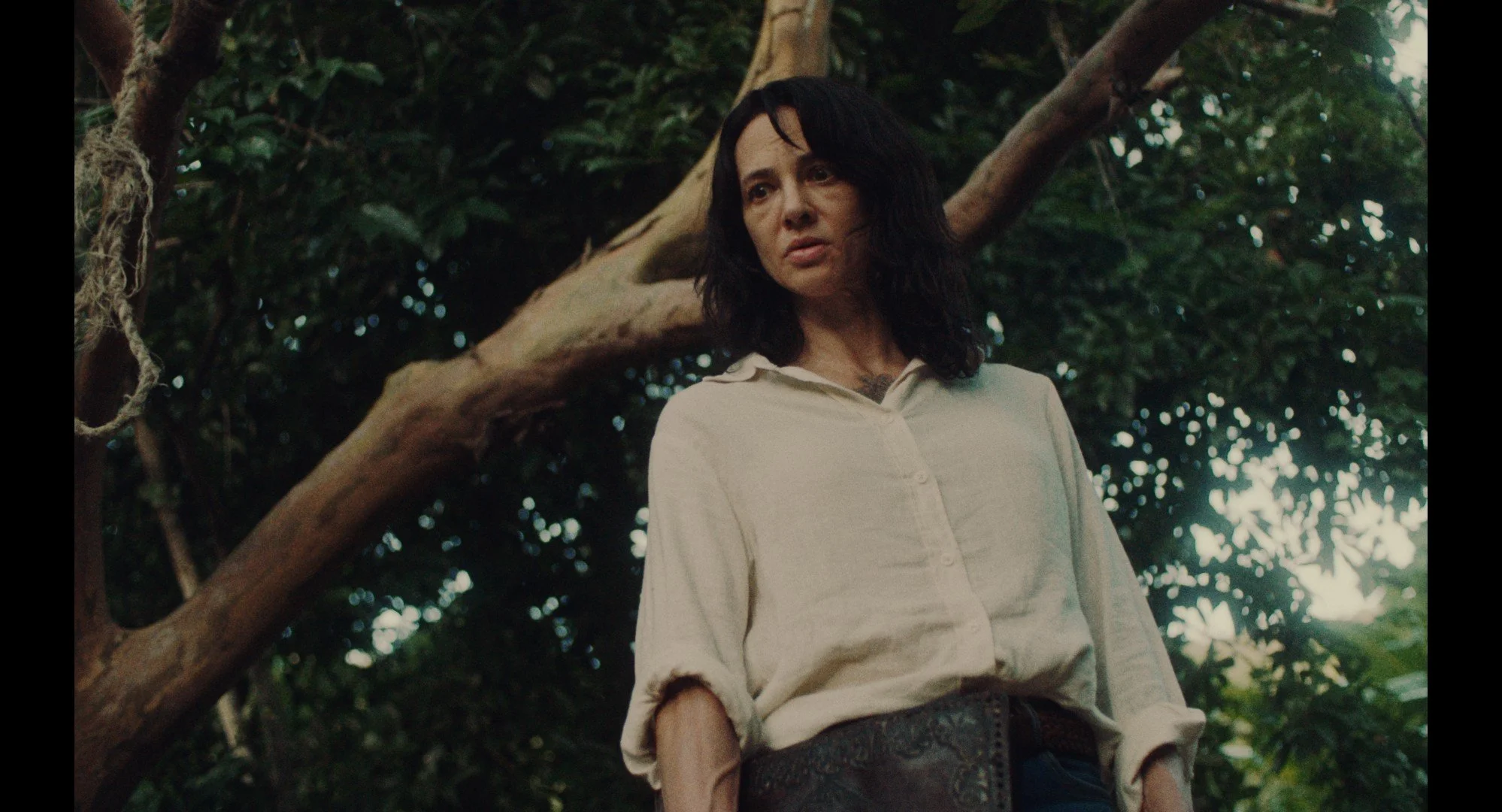 Asia Argento as Caro in Death Has No Master, standing in dense green jungle seen from a low angle, wearing a cream linen shirt, with a rope hanging from a tree branch beside her.