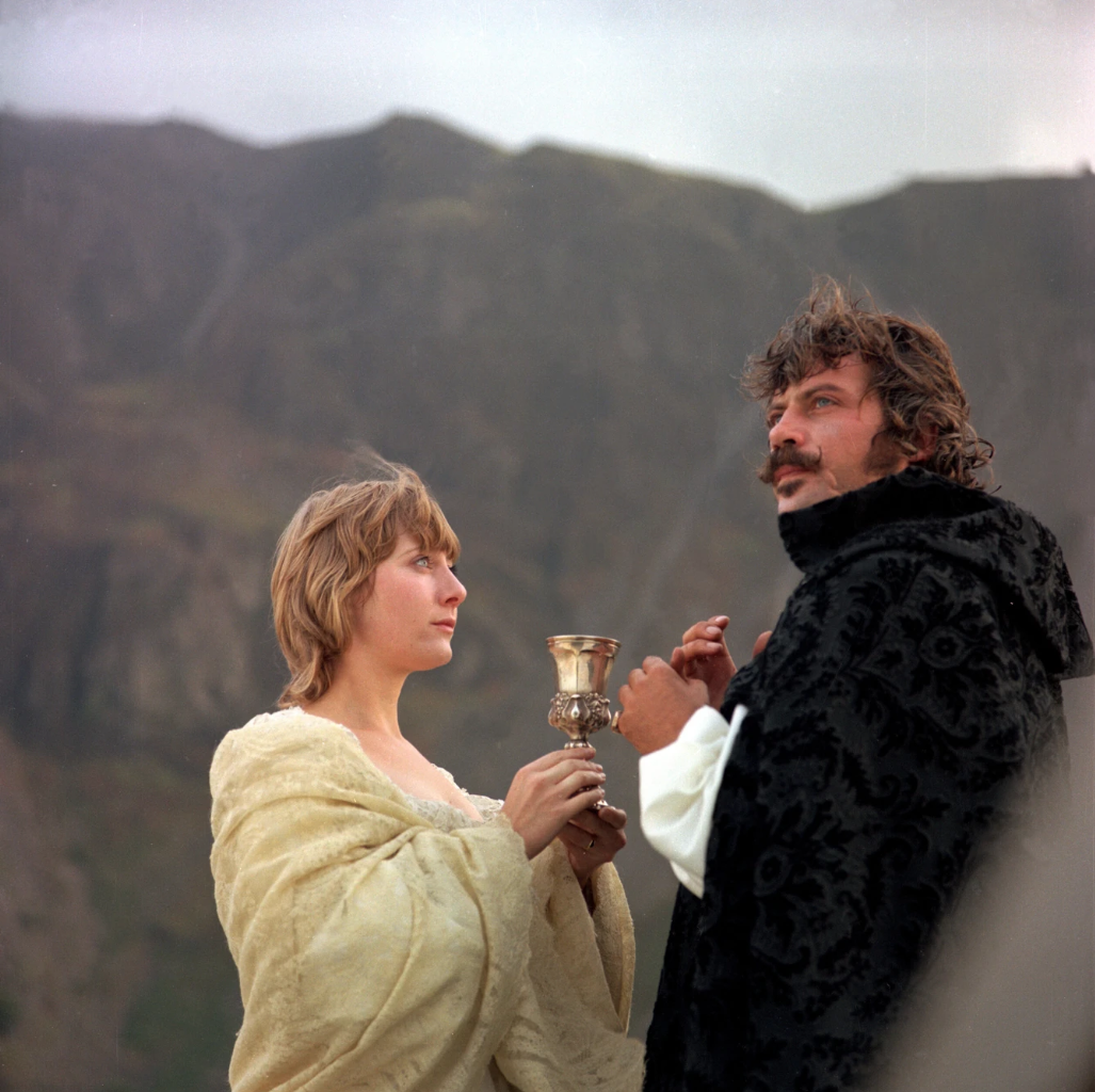 Oliver Reed as Father Urbain Grandier in black brocade, standing on a hillside beside a woman holding a silver chalice, in Ken Russell's The Devils (1971)