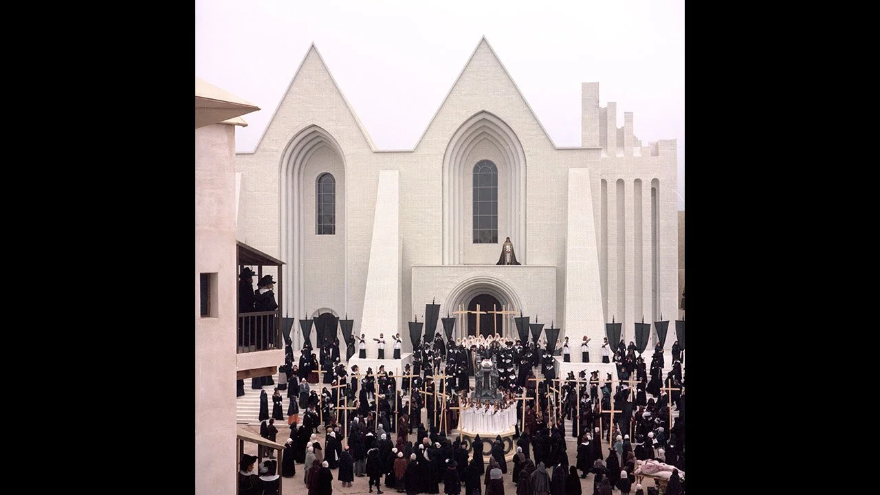 A crowd in black carrying wooden crosses before the enormous white cathedral set built for Ken Russell's The Devils (1971)