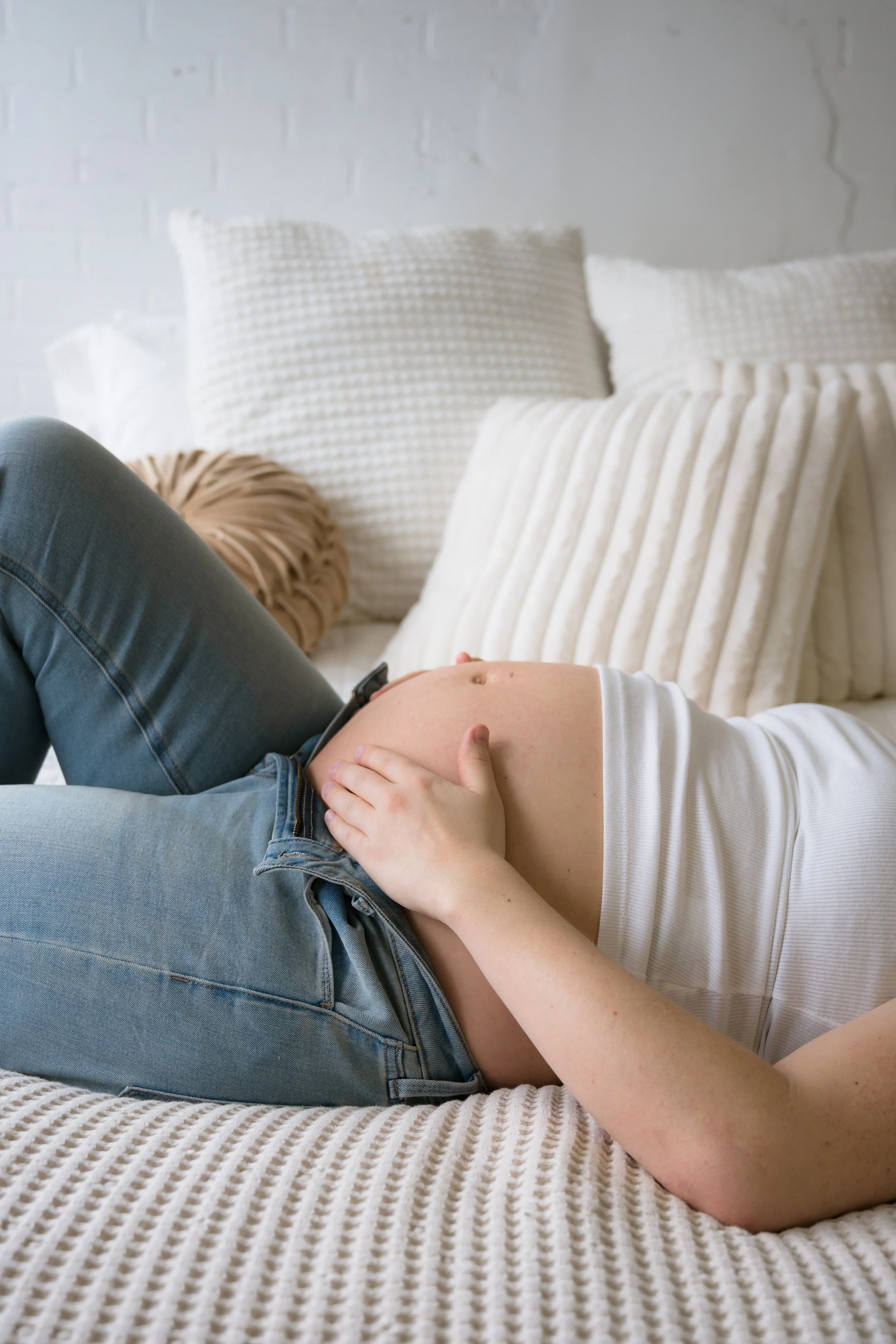 A pregnant woman lying on a bed with white textured pillows, resting her hand on her belly.
