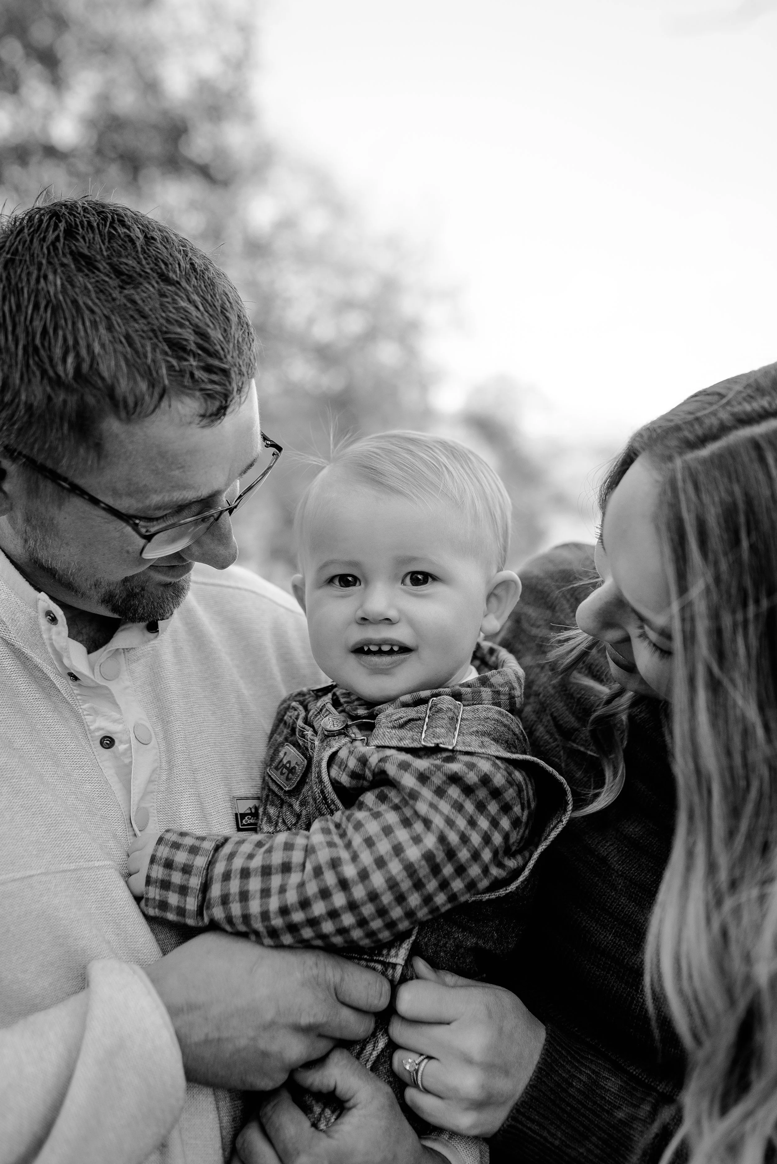 A black-and-white photo of a family outdoors, showing a man and a woman holding a young child between them, all smiling and looking at the camera.