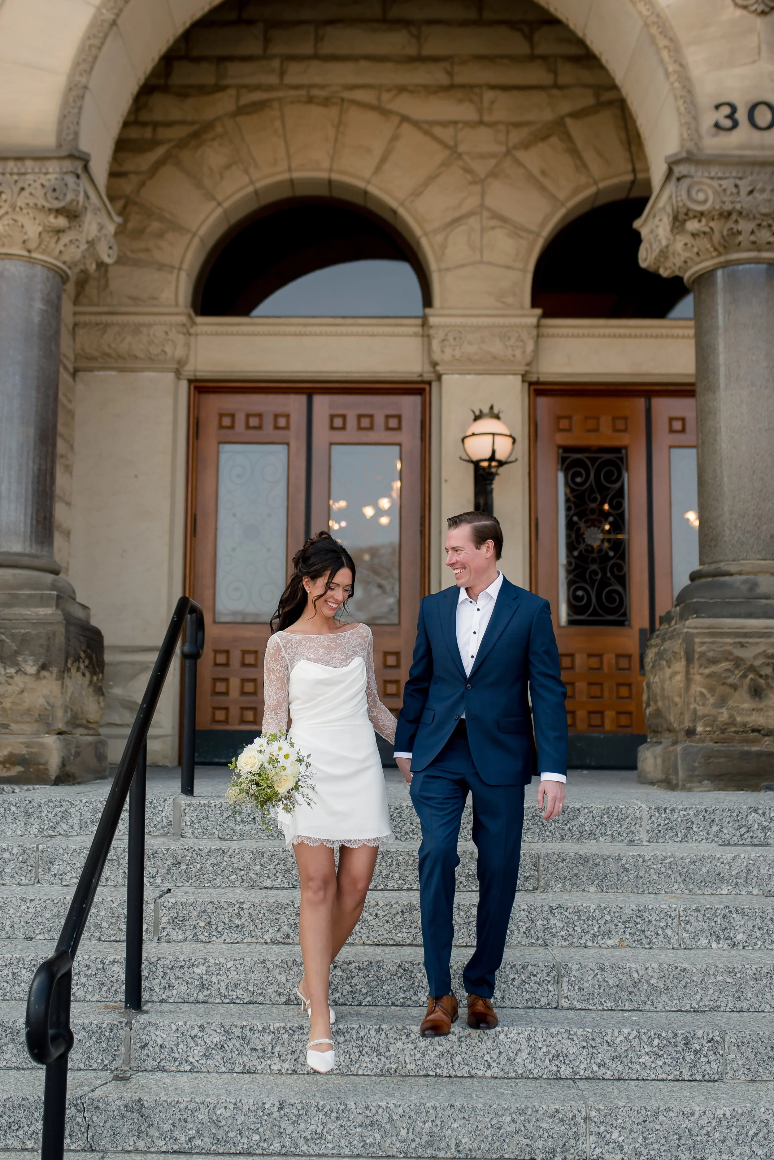 A newly married couple descending the steps outside a historic stone building, smiling and looking at each other. The woman is in a white lace cocktail dress and holding a bouquet, and the man is in a navy suit with brown shoes. The entrance features
