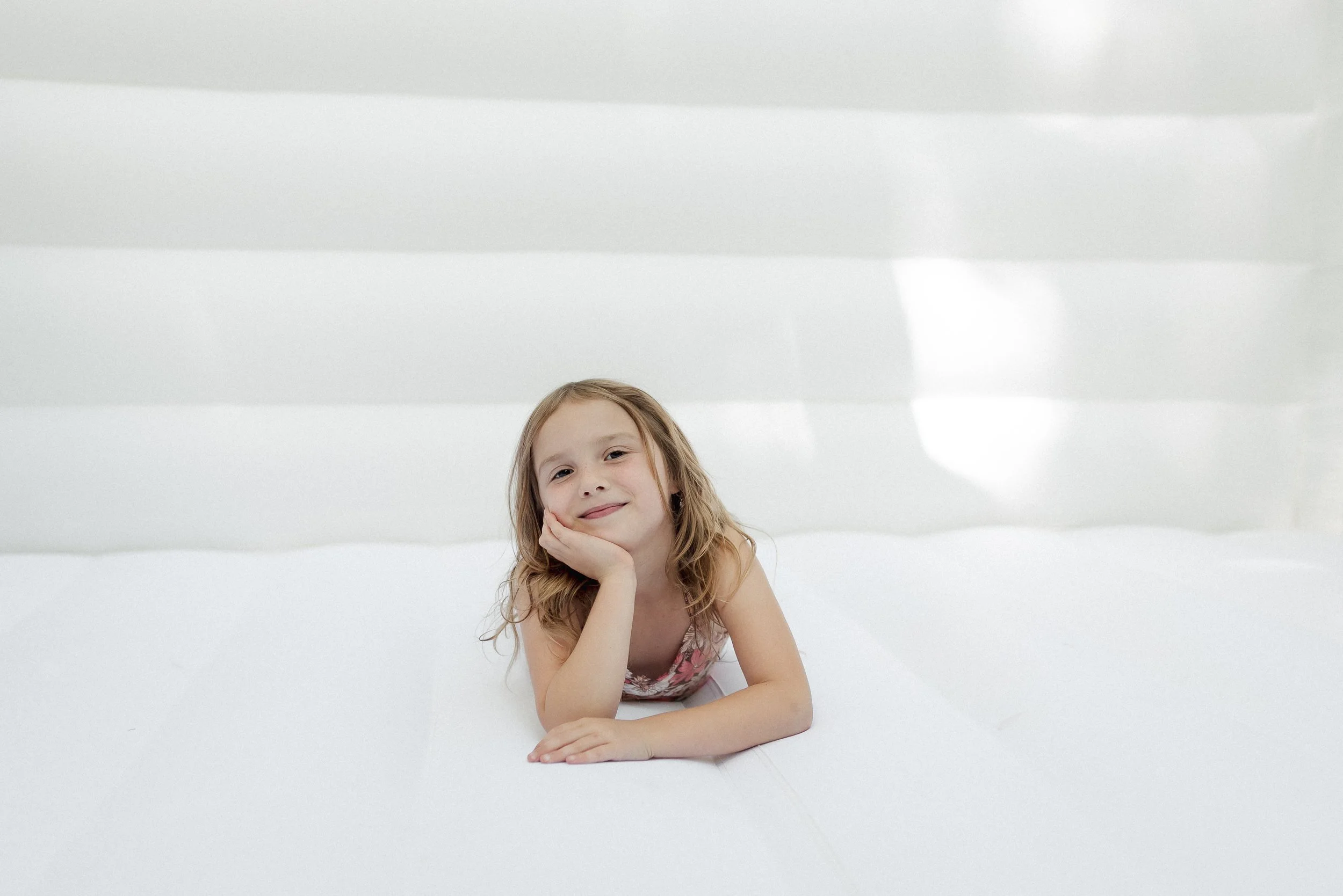 A young girl with blonde hair lying on a white bed with mylar blinds in the background, resting her chin on her hand and smiling at the camera.