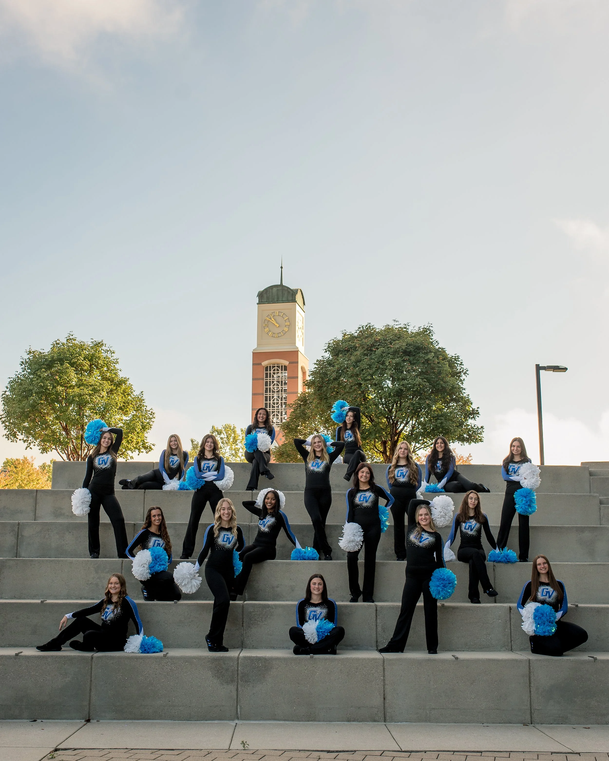 A cheerleading team in black and blue uniforms with blue and white pom-poms, posing on steps outdoors with a clock tower and trees in the background.