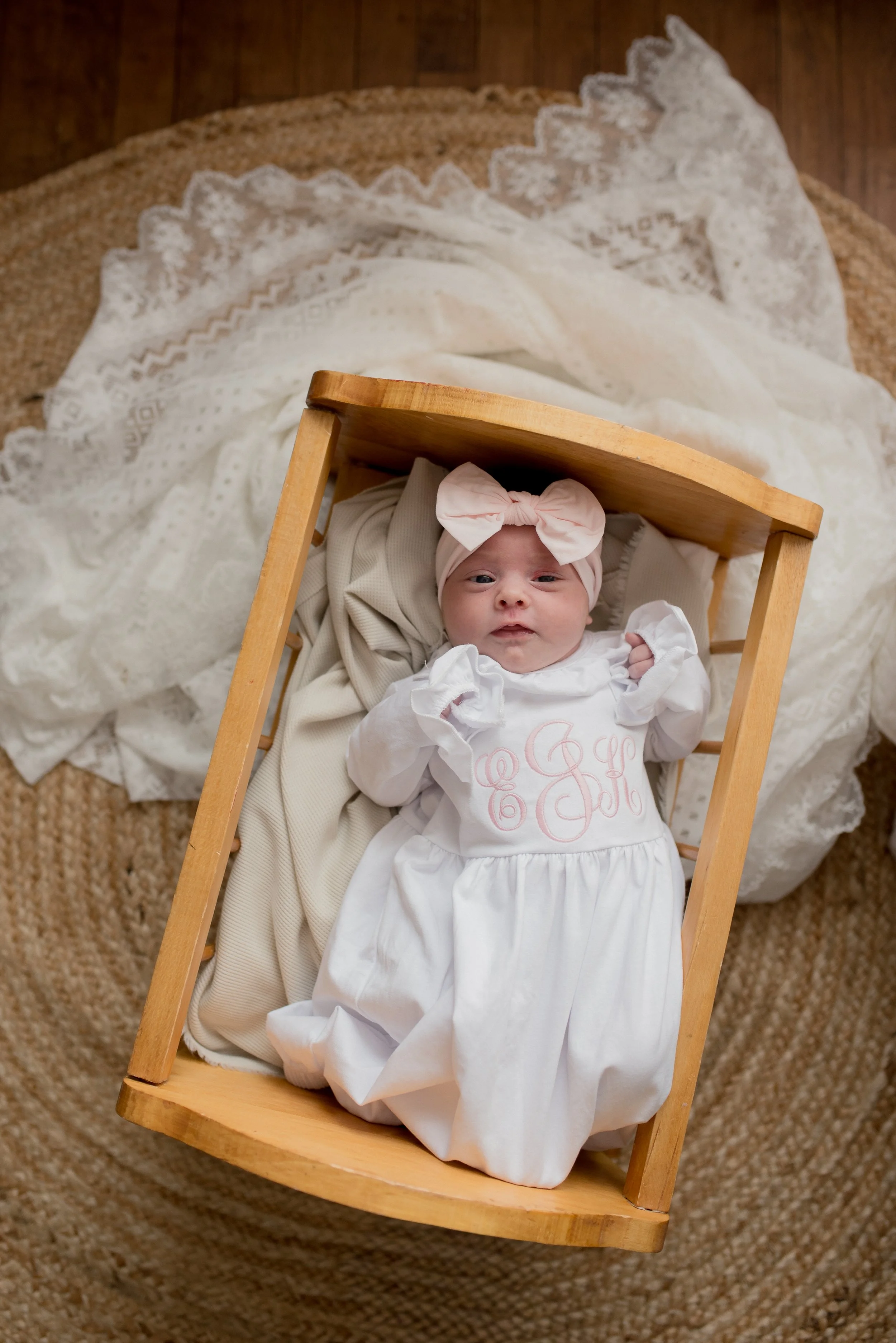 A baby girl lying in a wooden cradle on a woven rug, wearing a pink headband with a bow and a white dress with pink embroidery.