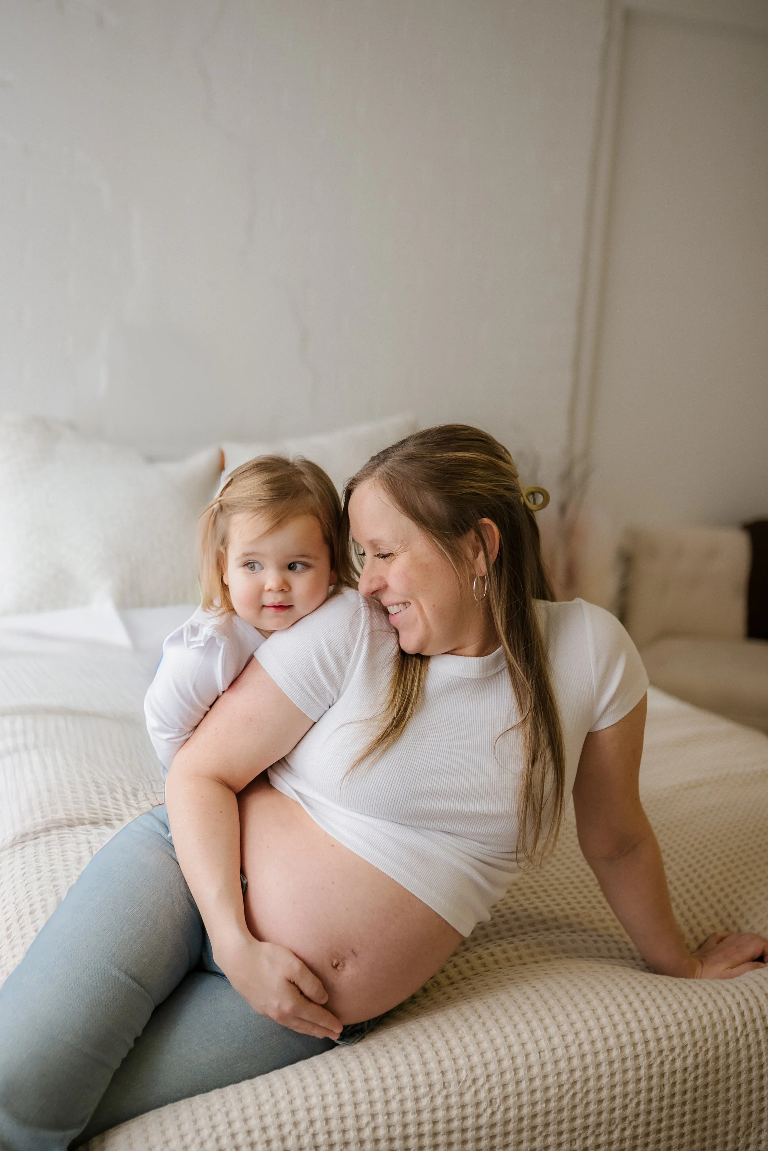 Mom and young girl on bed, mom's pregnant belly visible, mom smiling, girl resting her head on mom's shoulder, cozy bedroom setting