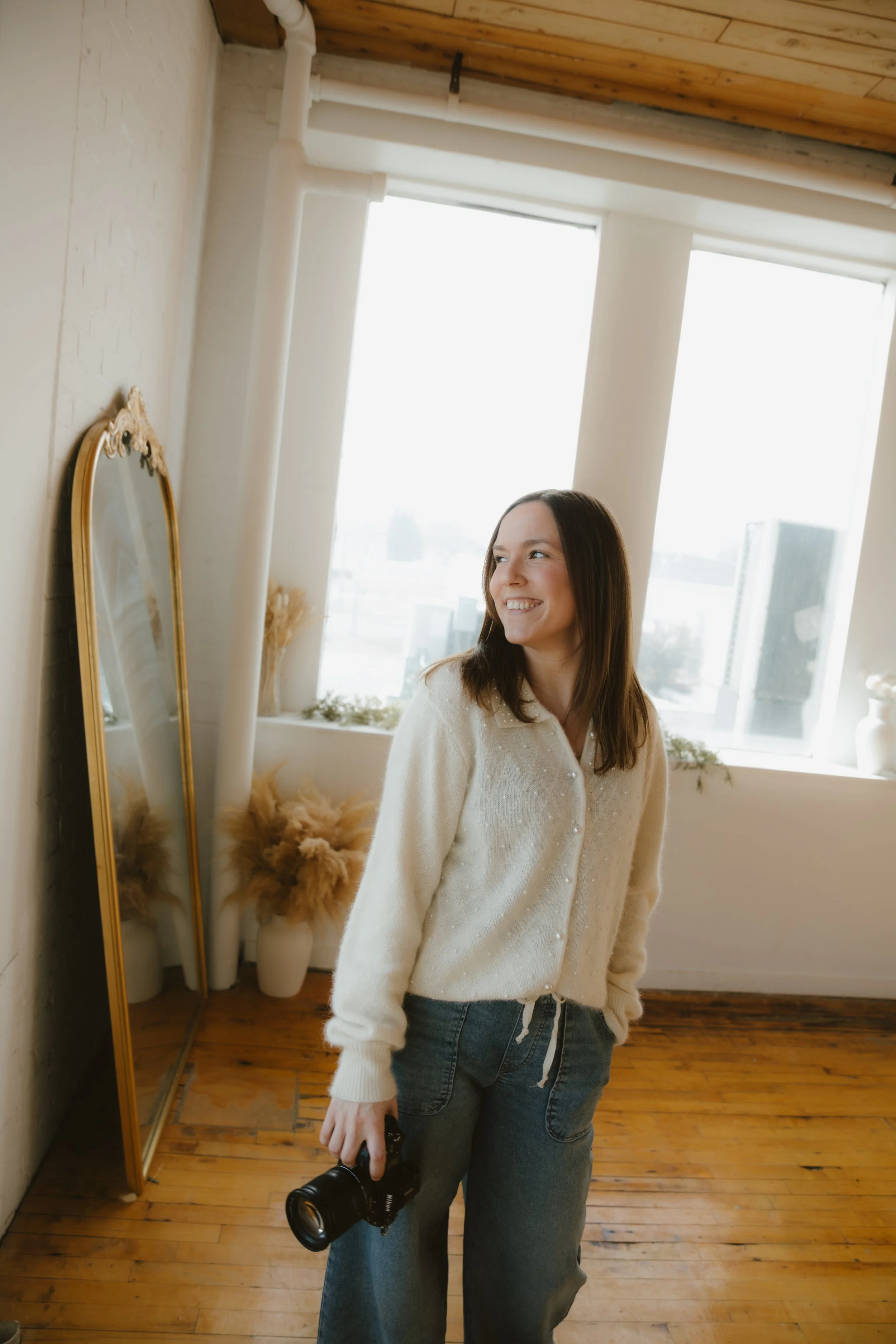 A young woman with shoulder-length dark hair and light skin smiling, holding a black camera in her right hand, indoors near a large window with white walls, wooden floor, and a vintage gold-framed mirror leaning against the wall.
