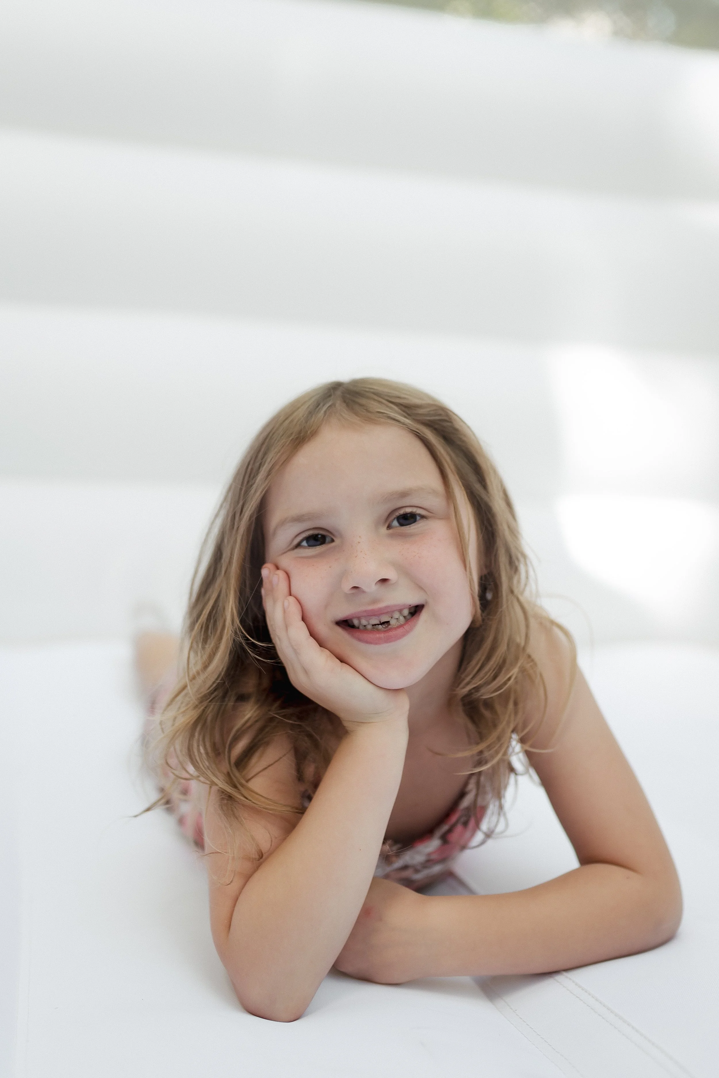 A young girl with red hair and freckles lying on her side, resting her face in her hand, smiling at the camera, with a white background.