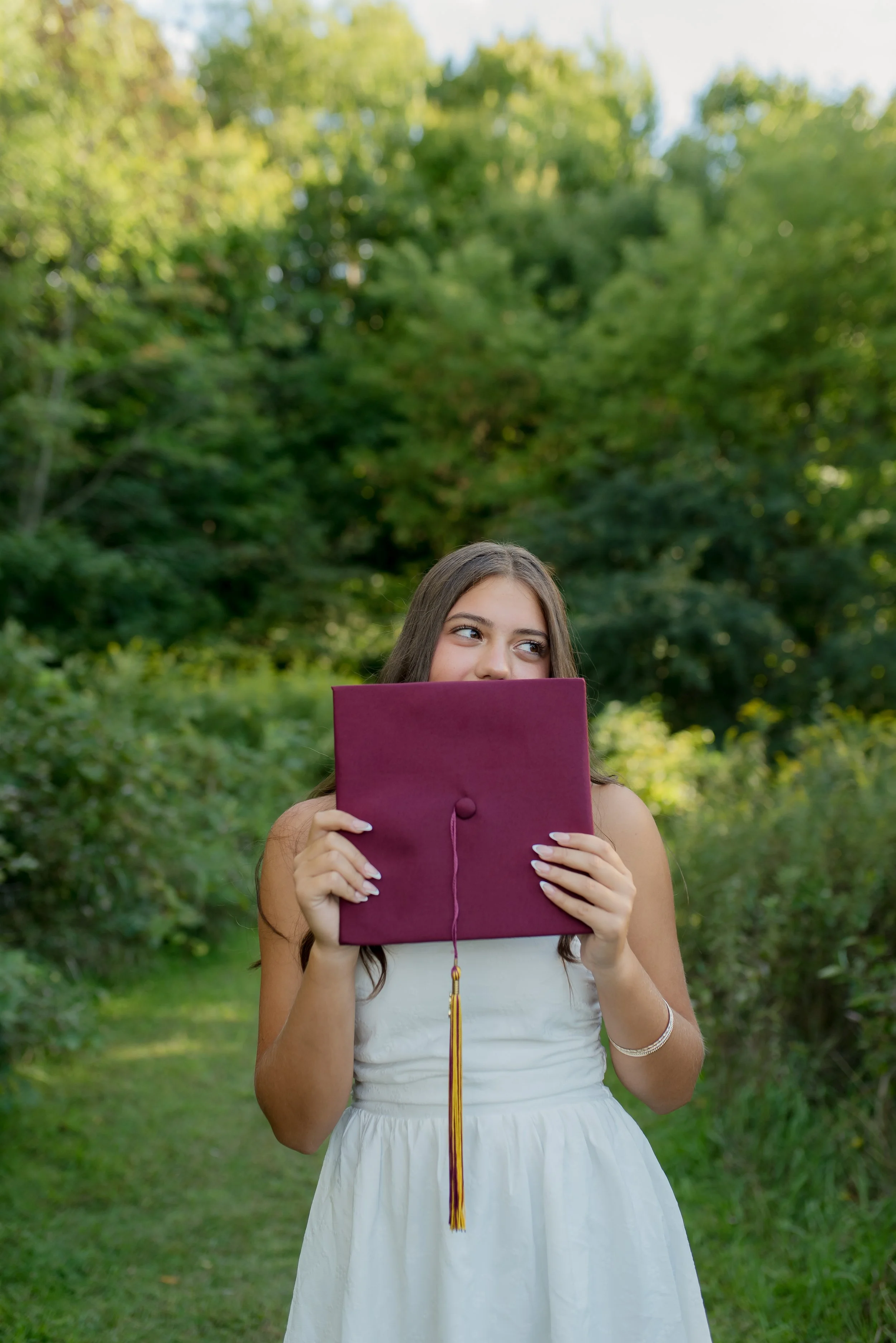 Young woman holding diploma with a purple cover, standing outdoors in front of green trees, dressed in a white dress, looking to the side.
