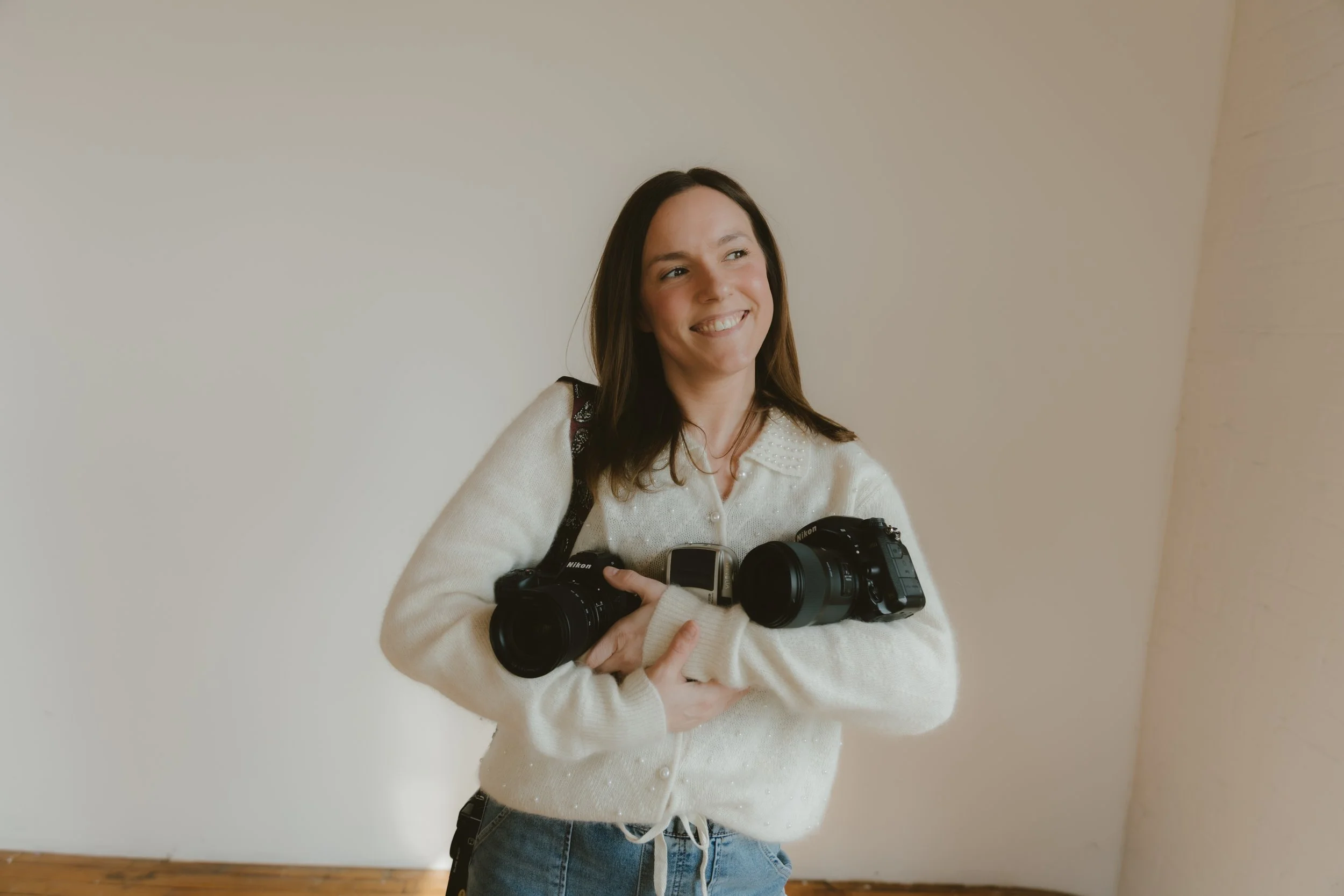 A young woman holding two cameras, smiling, standing in a room with a white wall and wooden floor.