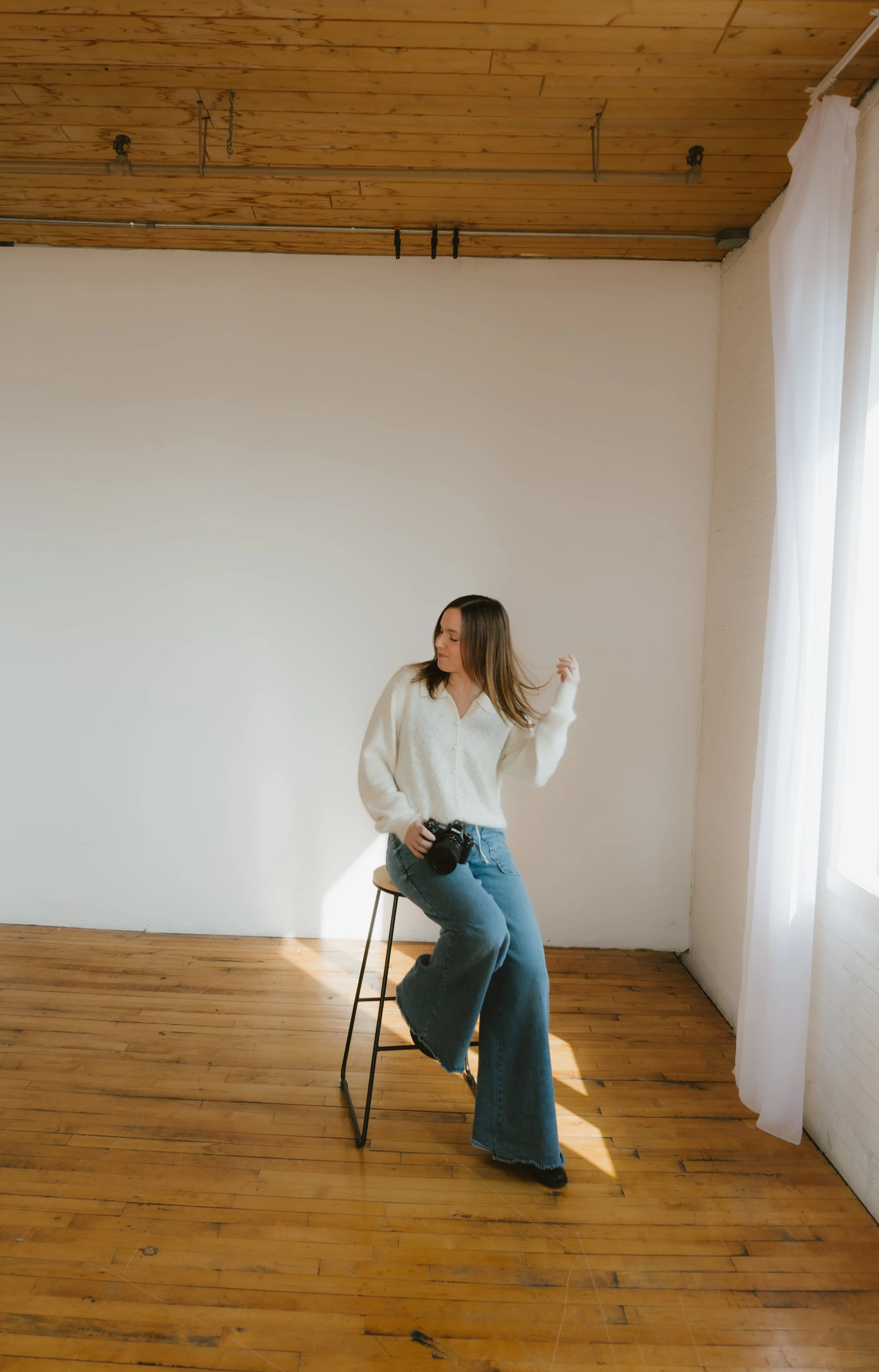 A woman sitting on a stool in a bright room with wooden floors and ceiling, holding a camera in her lap, with white curtains and sunlight coming in from the right.