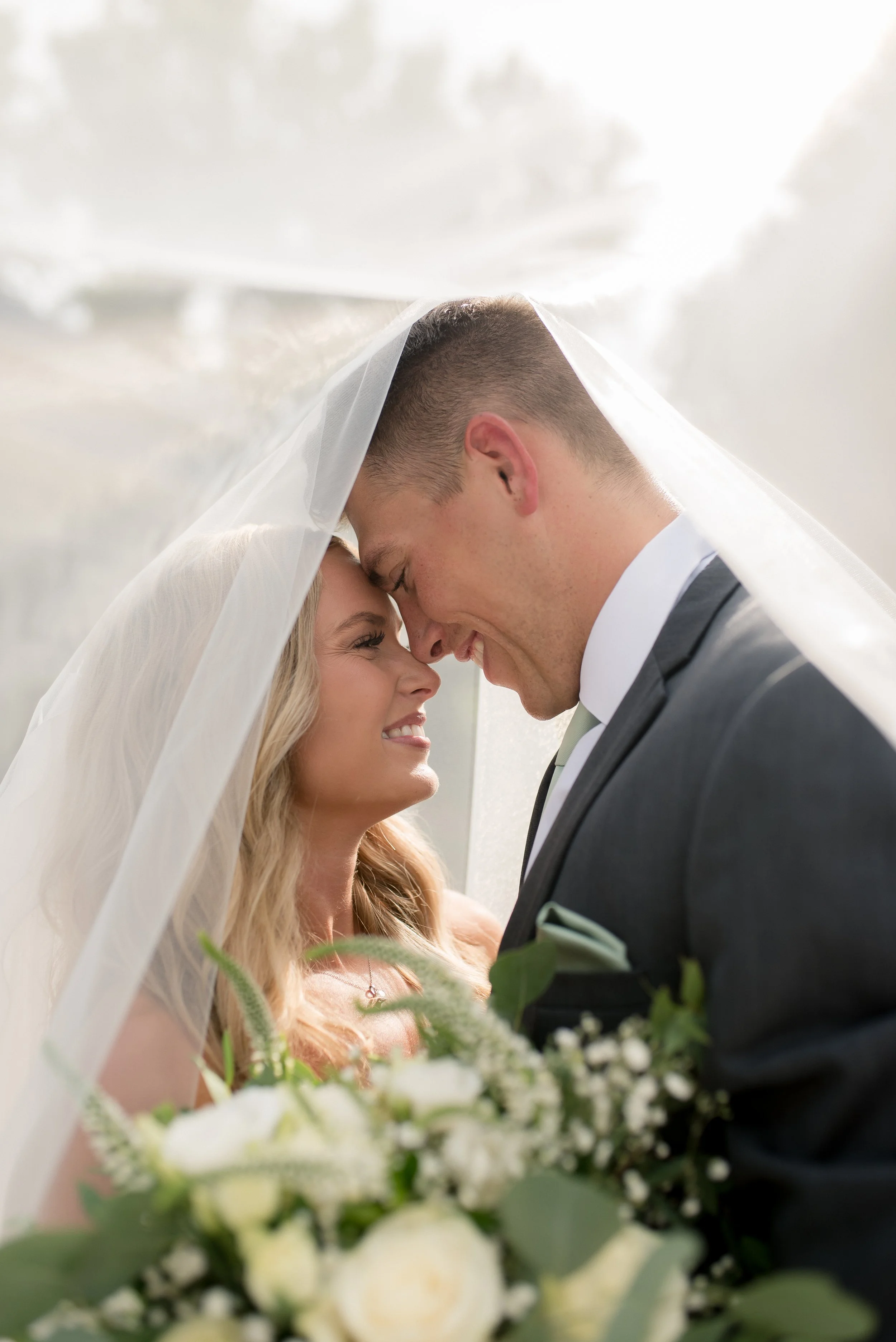 A bride and groom touching foreheads and smiling beneath a wedding veil, with a bouquet of white roses and greenery in the foreground.
