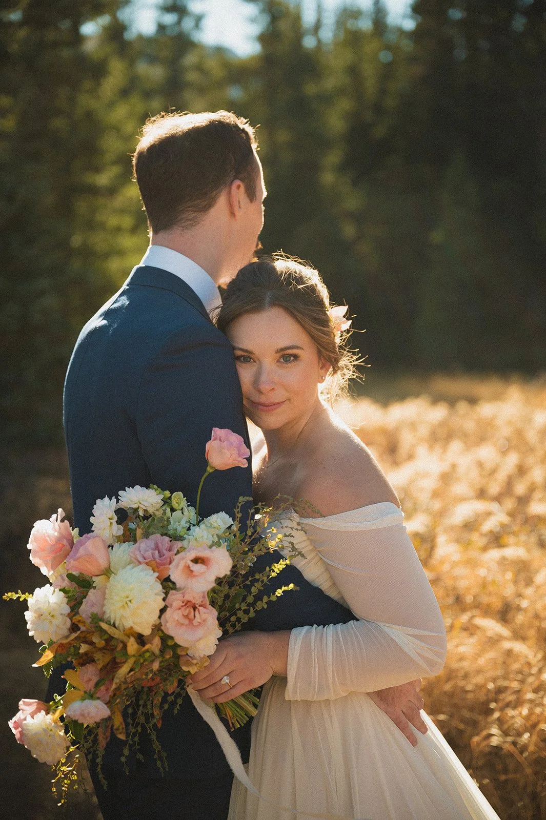 A bride holding a flower bouquet looking at the camera and hugging her groom