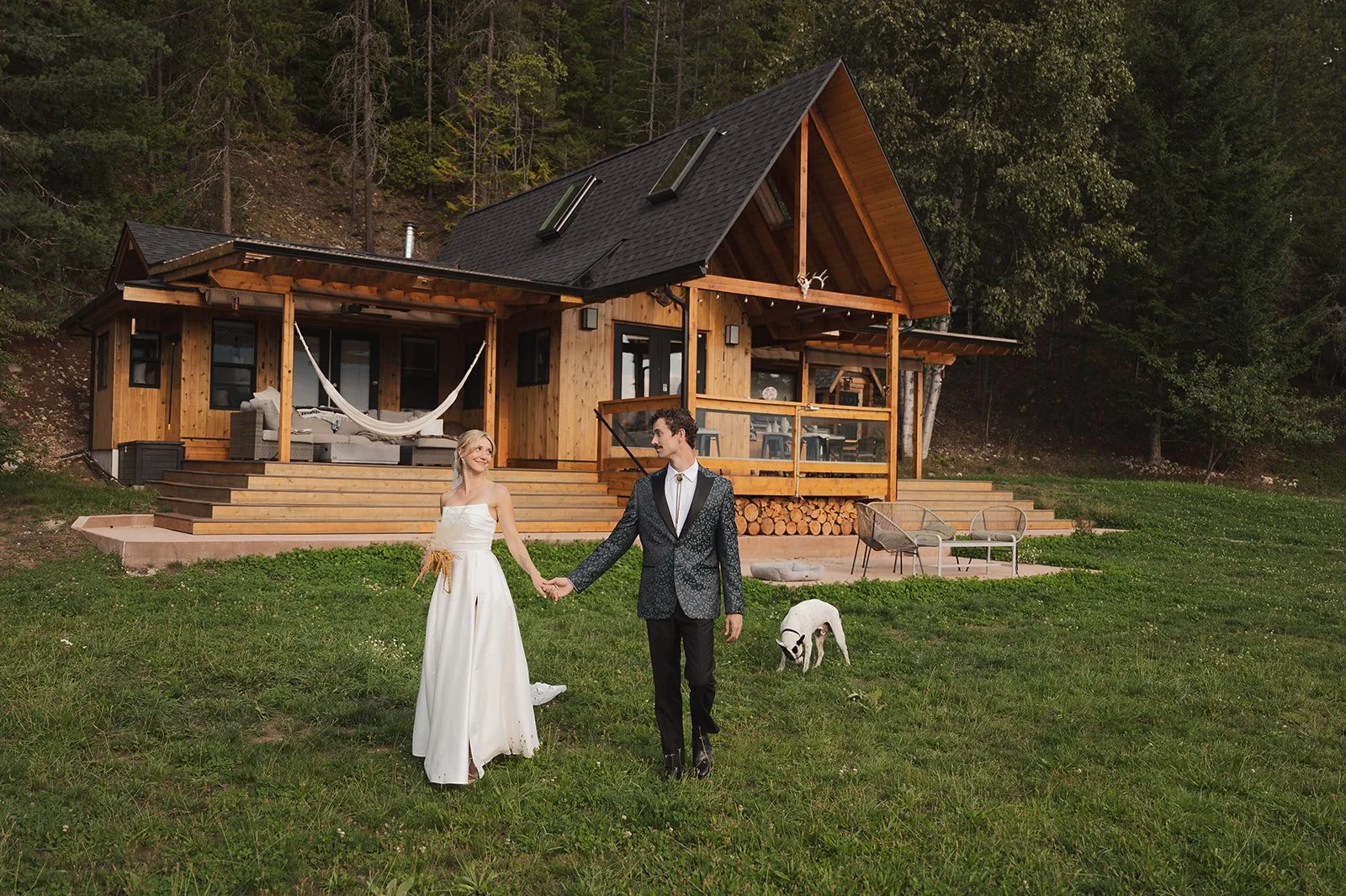 A bride and groom are celebrating getting married at an Airbnb, they are strolling hand in hand on a green lawn in front of a modern log cabin vacation rental