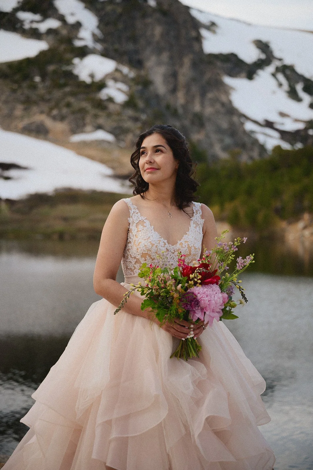 A bride in a lace and tulle wedding dress softly smiling and holding a bouquet of wildflowers