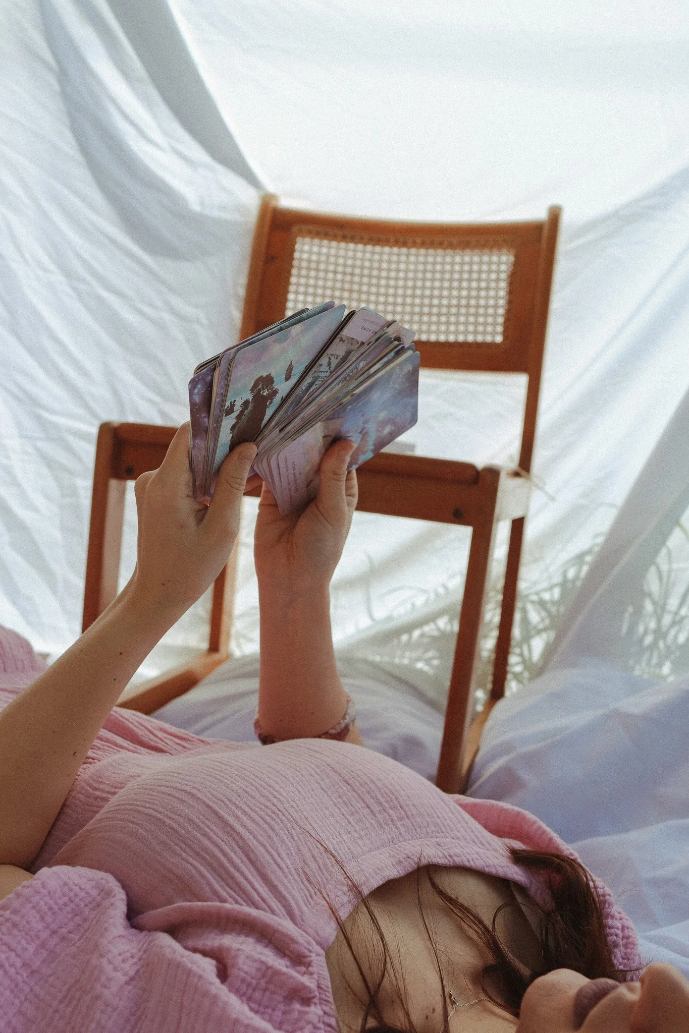 Person lying on a bed holding tarot cards with a wooden chair and white sheet in the background.