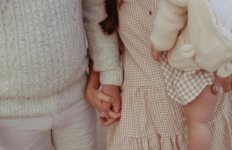 Close-up of a family holding hands, wearing cozy knit sweaters and a checkered dress. A baby is partially visible, held by one adult.
