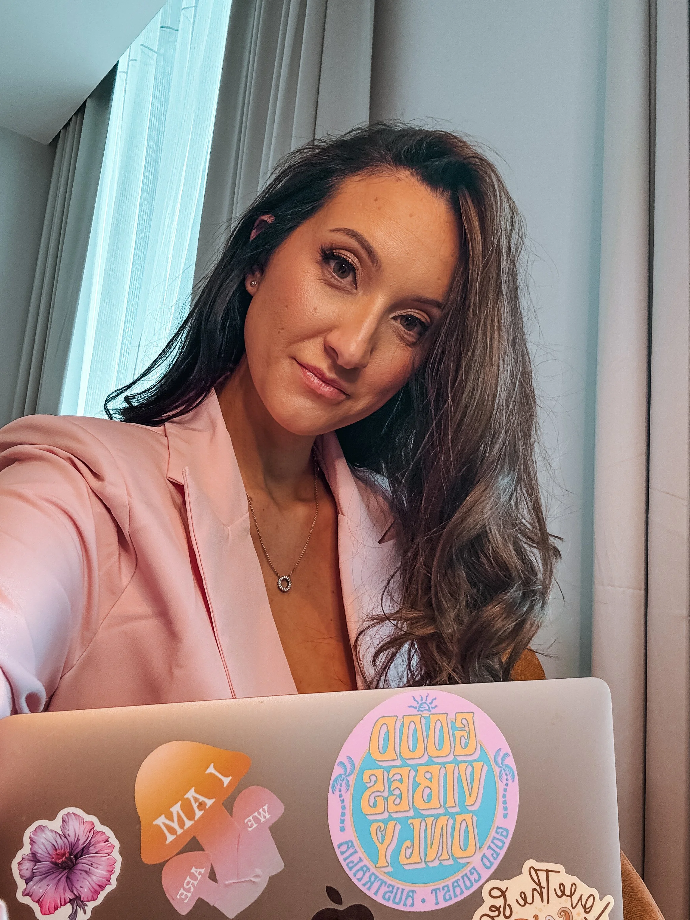 A woman with long wavy brown hair wearing a light pink blazer, sitting near a window with sheer curtains. She is smiling slightly and looking into the camera, with stickers on her laptop, including a colorful