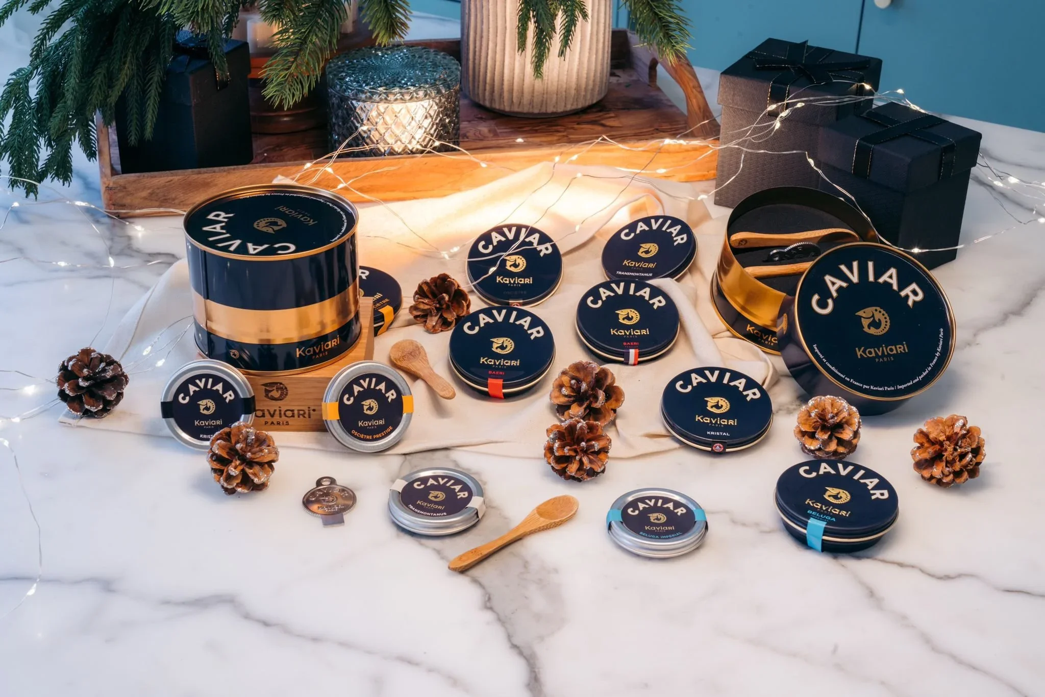 Arrangement of Kaviari brand caviar tins on a marble table, decorated with pinecones, wooden spoons, and fairy lights, with a wooden backdrop and Christmas tree branches overhead.