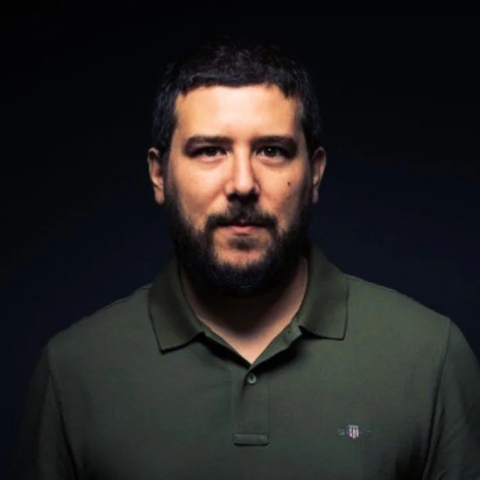Man in a dark green polo shirt with a beard, facing forward against a dark background.