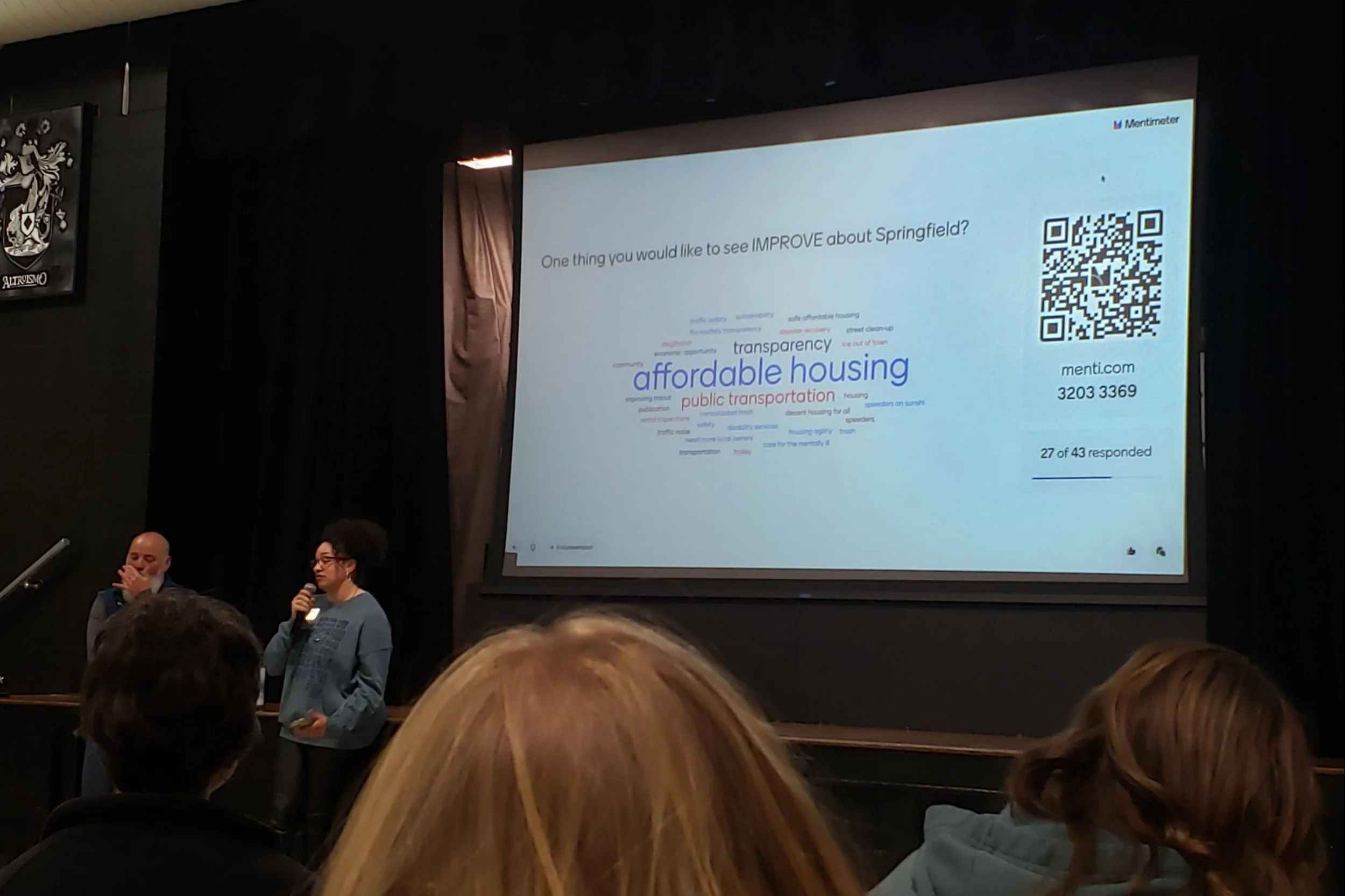 Springfield City Council members Bruce Adib-Yazdi and Heather Hardinger stand in front of a town hall audience. Behind them is a large projector screen showing a QR code and many different submitted keywords from audience members.