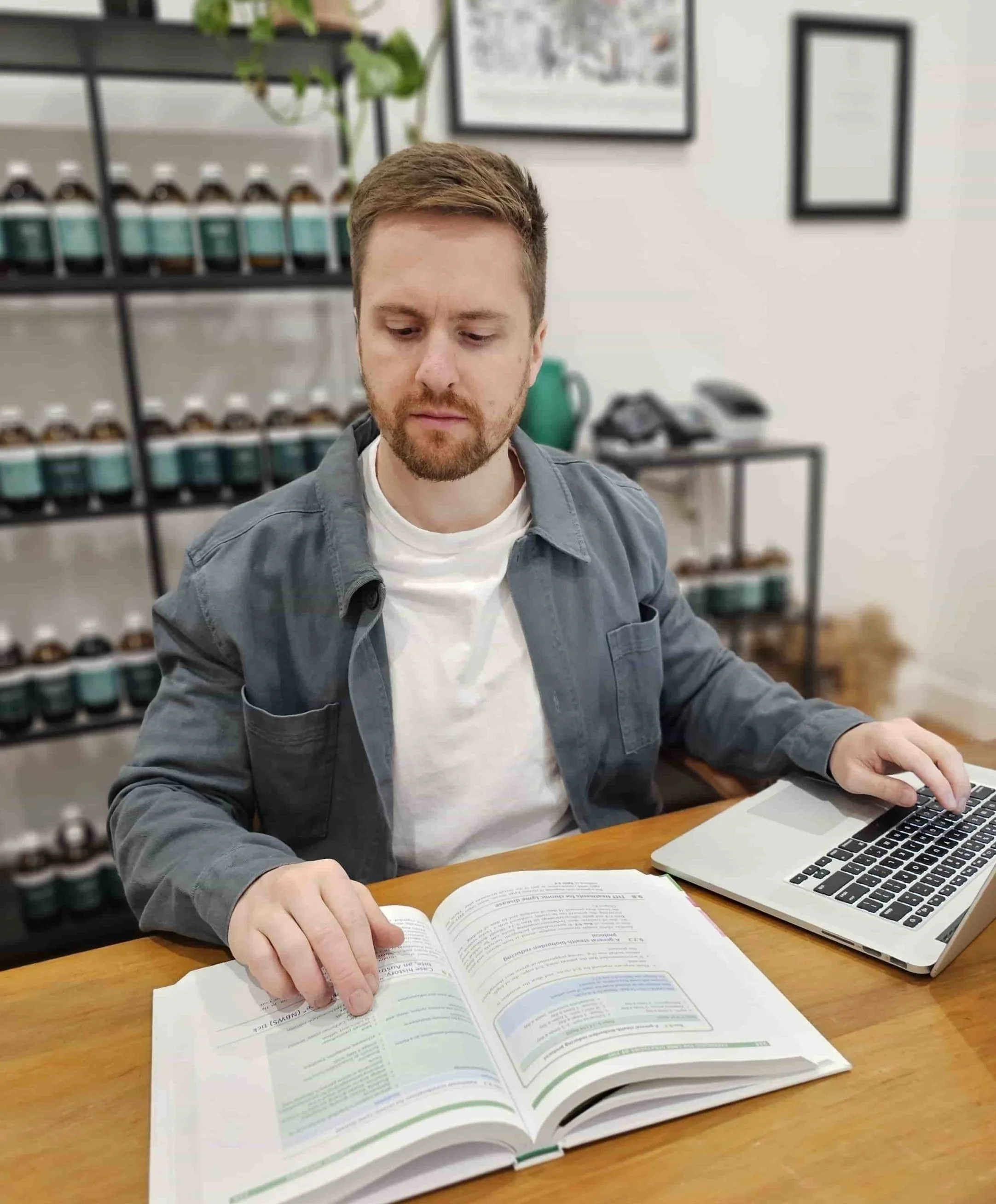 A man sitting at a wooden table with an open book and a laptop, in a room with shelves of jars in the background, looking at the book.