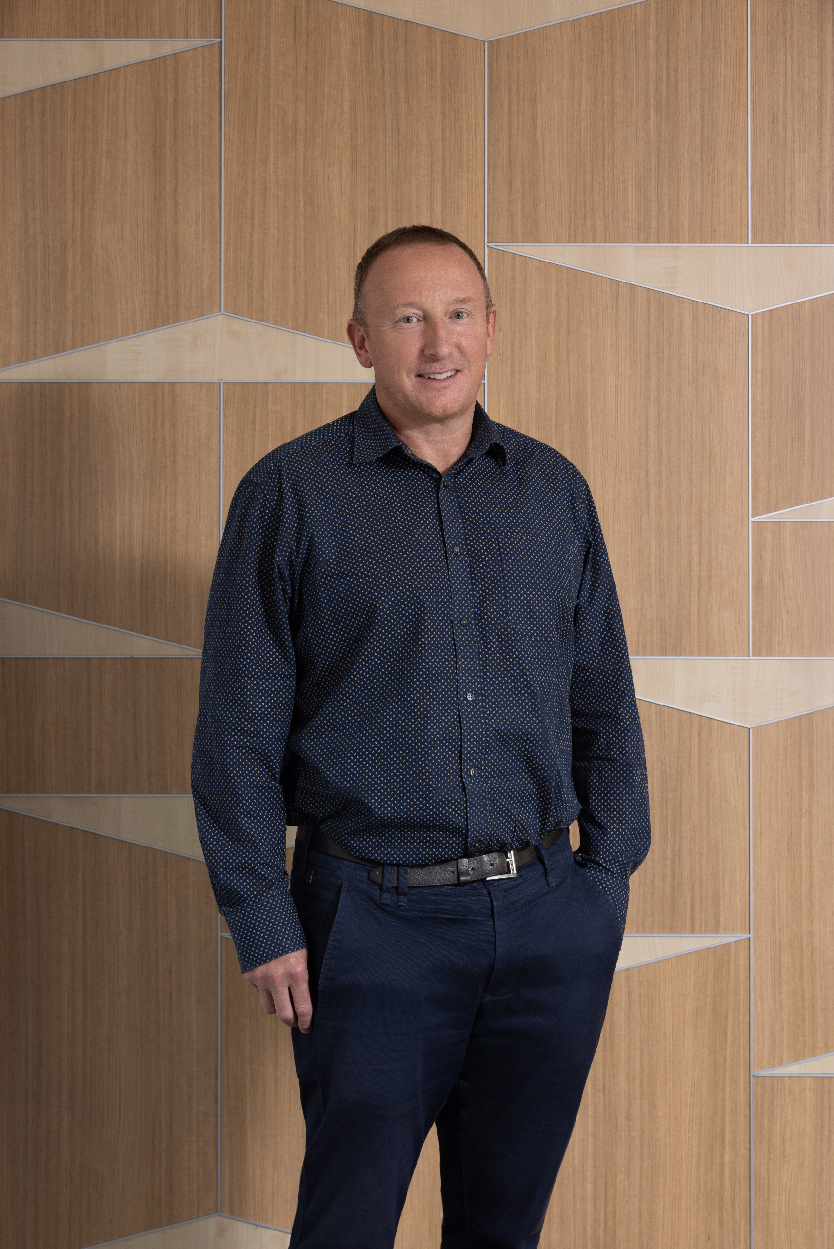 A man standing against a wooden wall with geometric panels, wearing a dark blue shirt with small white dots and dark blue pants.
