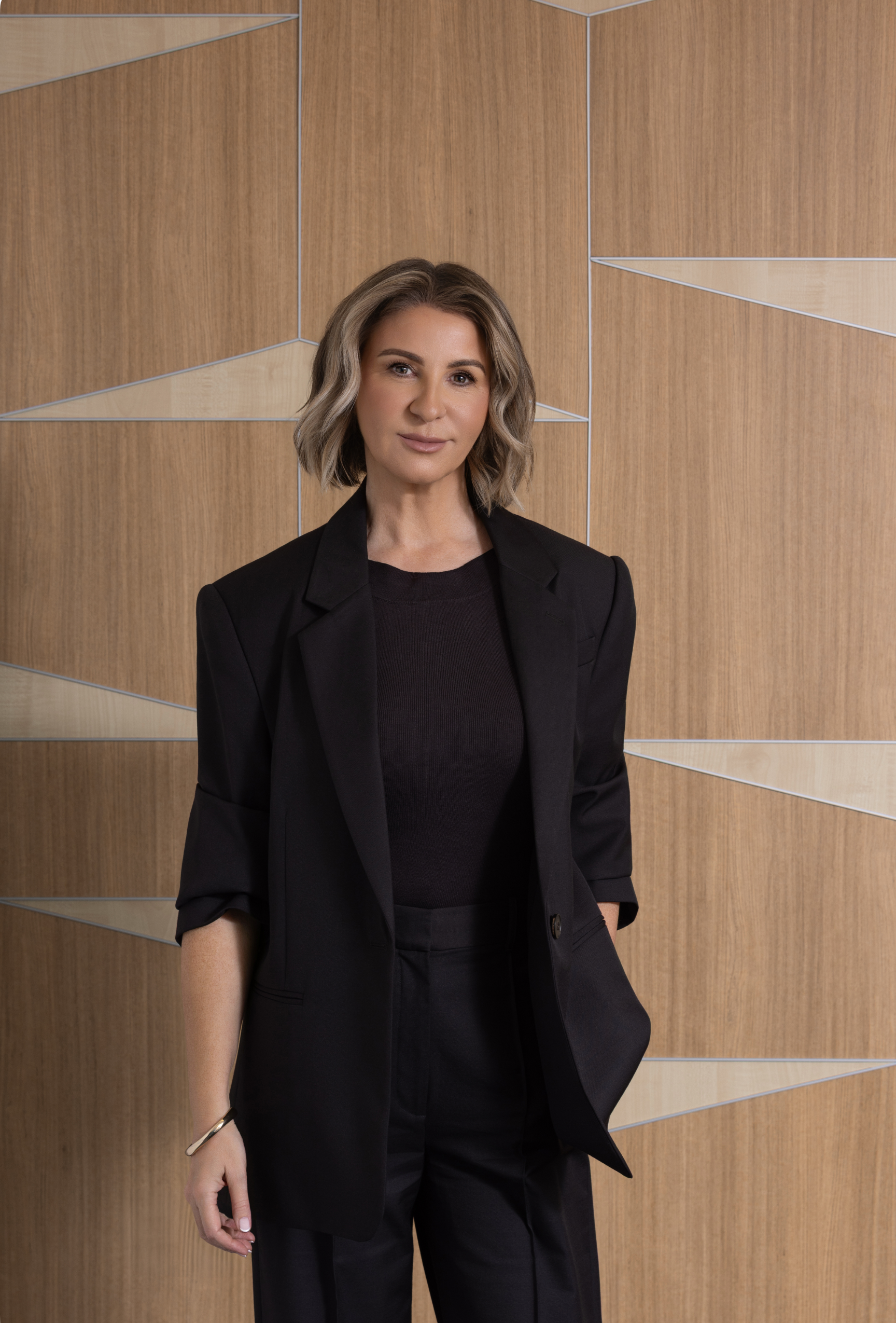 Woman with blond hair wearing a black blazer and white shirt, standing in front of a geometric wooden wall, smiling slightly.