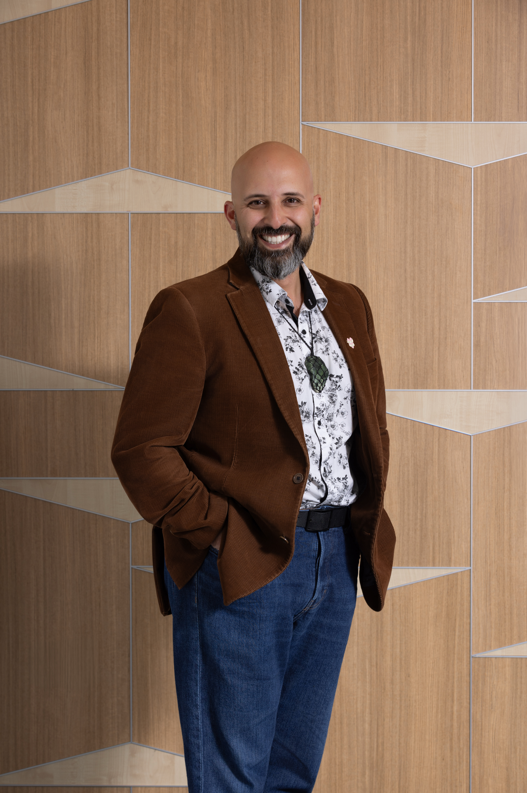 A smiling man with a bald head, beard, and mustache, wearing a brown blazer, a patterned shirt, and blue jeans, standing in front of a wooden wall with geometric patterns.