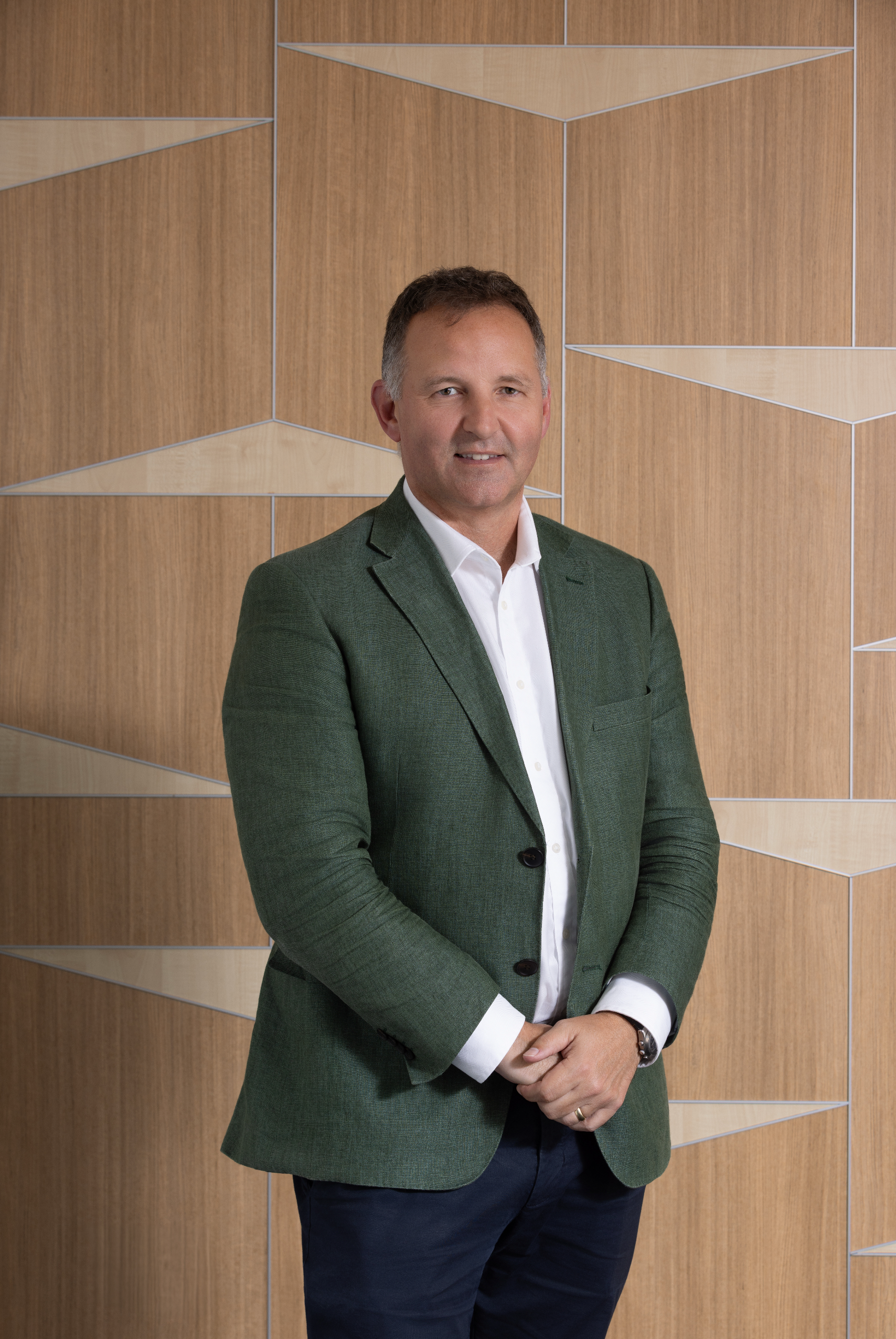 Man in a plaid shirt and blue blazer smiling, standing against a geometric wooden wall.