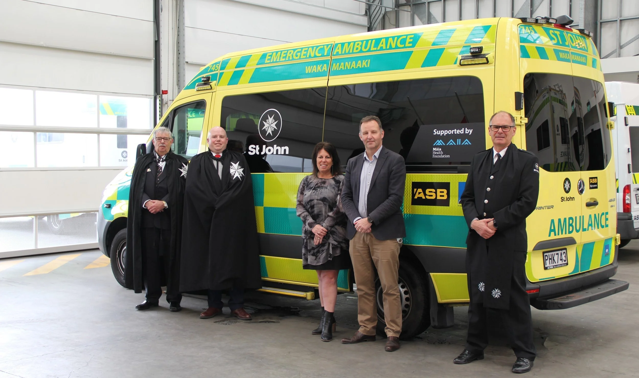 A group of five people standing in front of a yellow and green St John ambulance inside a garage. Two are wearing ceremonial robes and three in business attire.